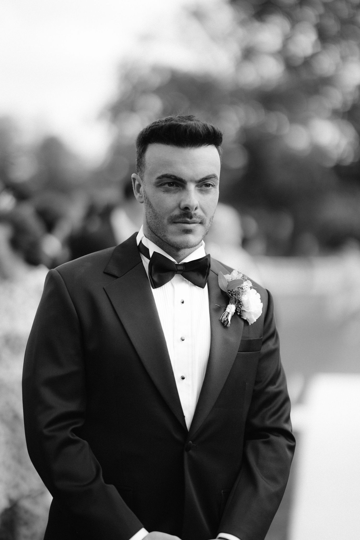 Elegant groom in black tuxedo with bow tie and boutonniere poses outdoors at wedding ceremony.