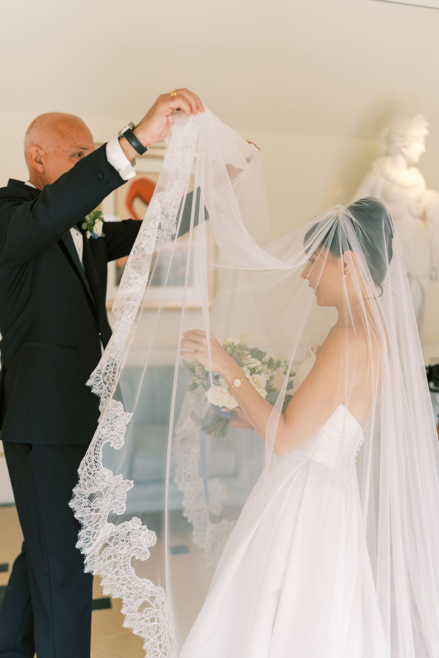A bald man in a tuxedo lifts a lace-trimmed wedding veil over a bride in a white gown during wedding preparations.