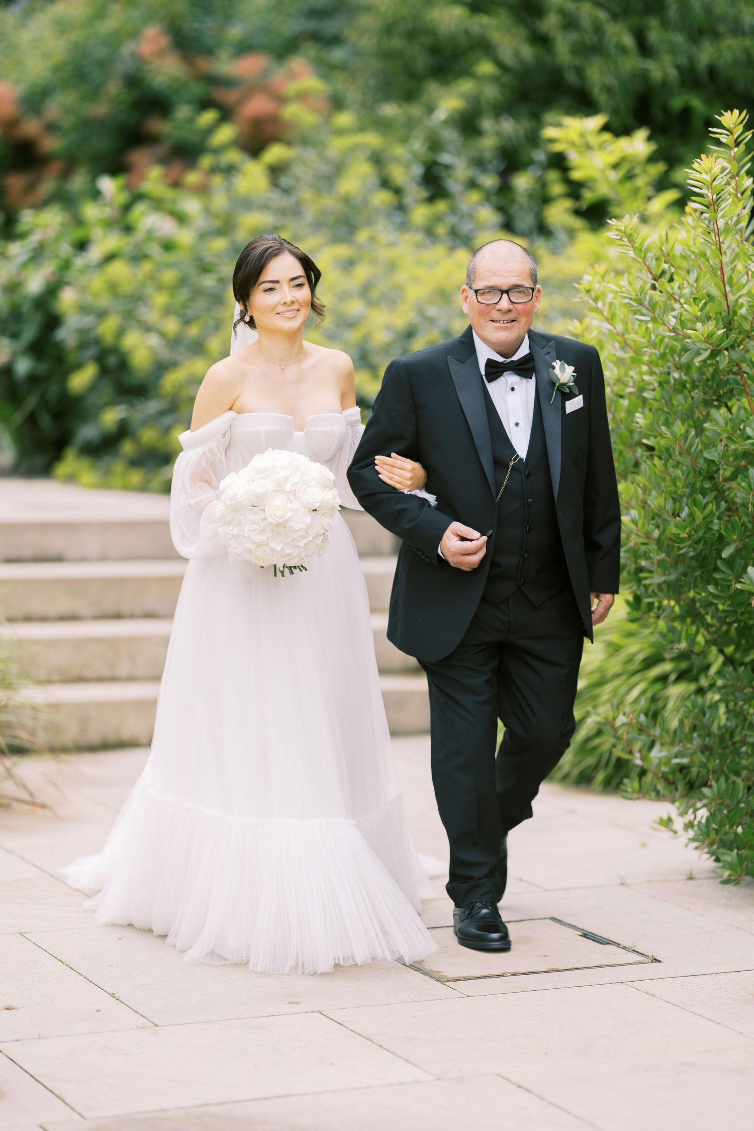 Bride in off-shoulder gown holding white bouquet walks arm-in-arm with father in tuxedo through garden path.