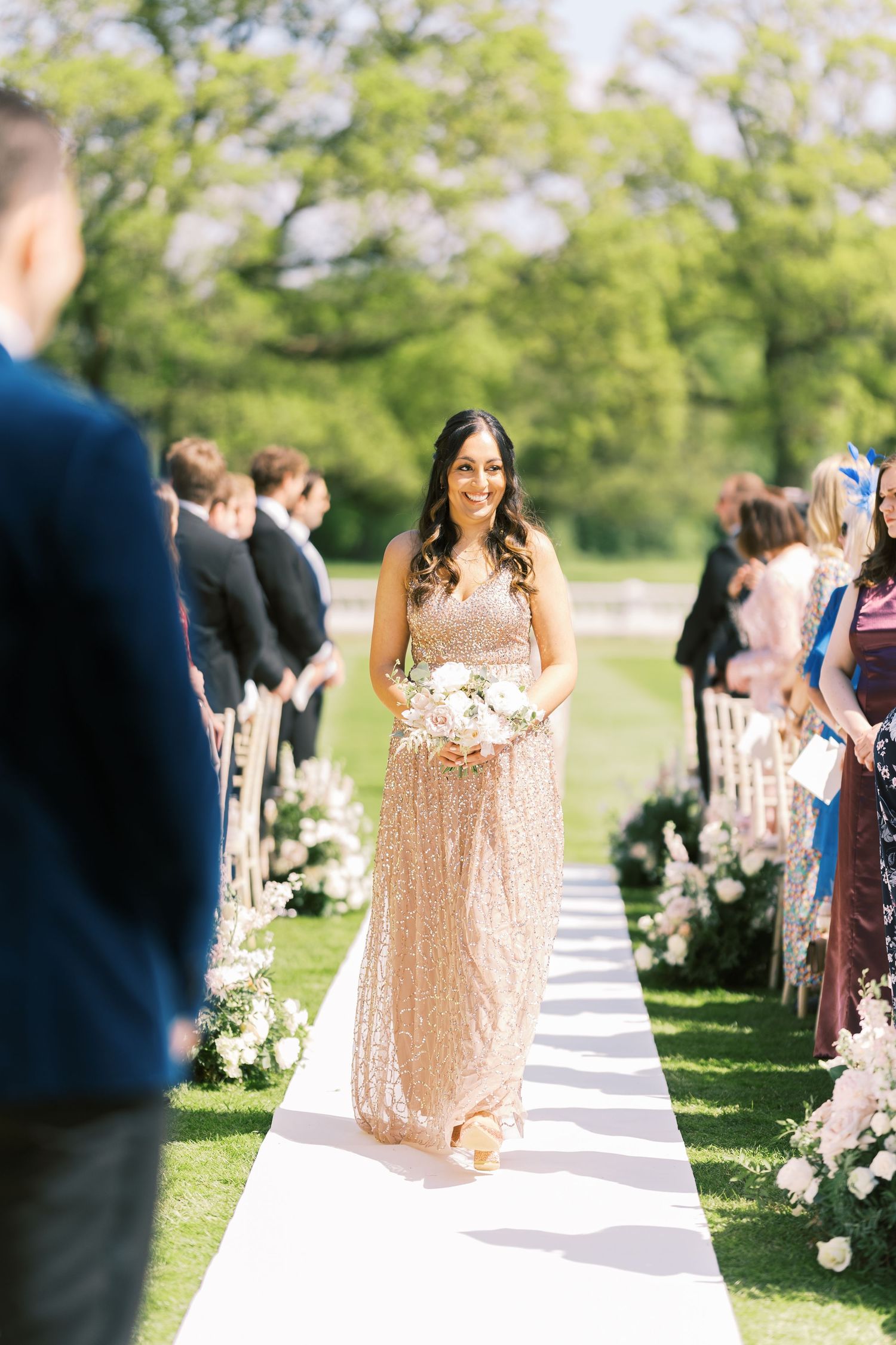 Bridesmaid in gold sequin gown walking down floral wedding aisle.