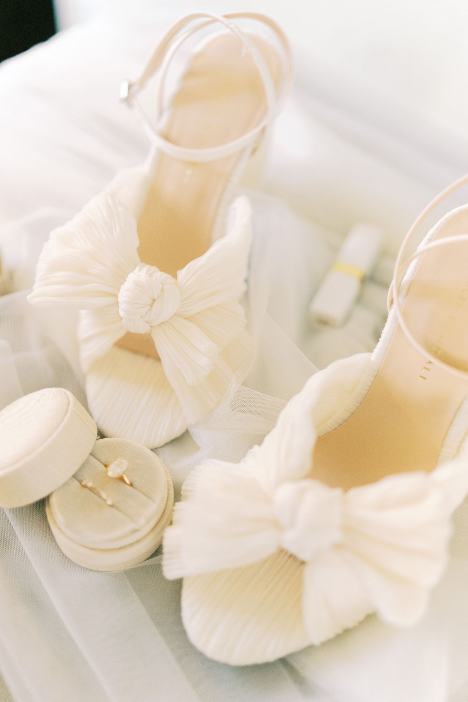 Elegant white bridal heels with bow detail beside a ring box on a soft white surface.