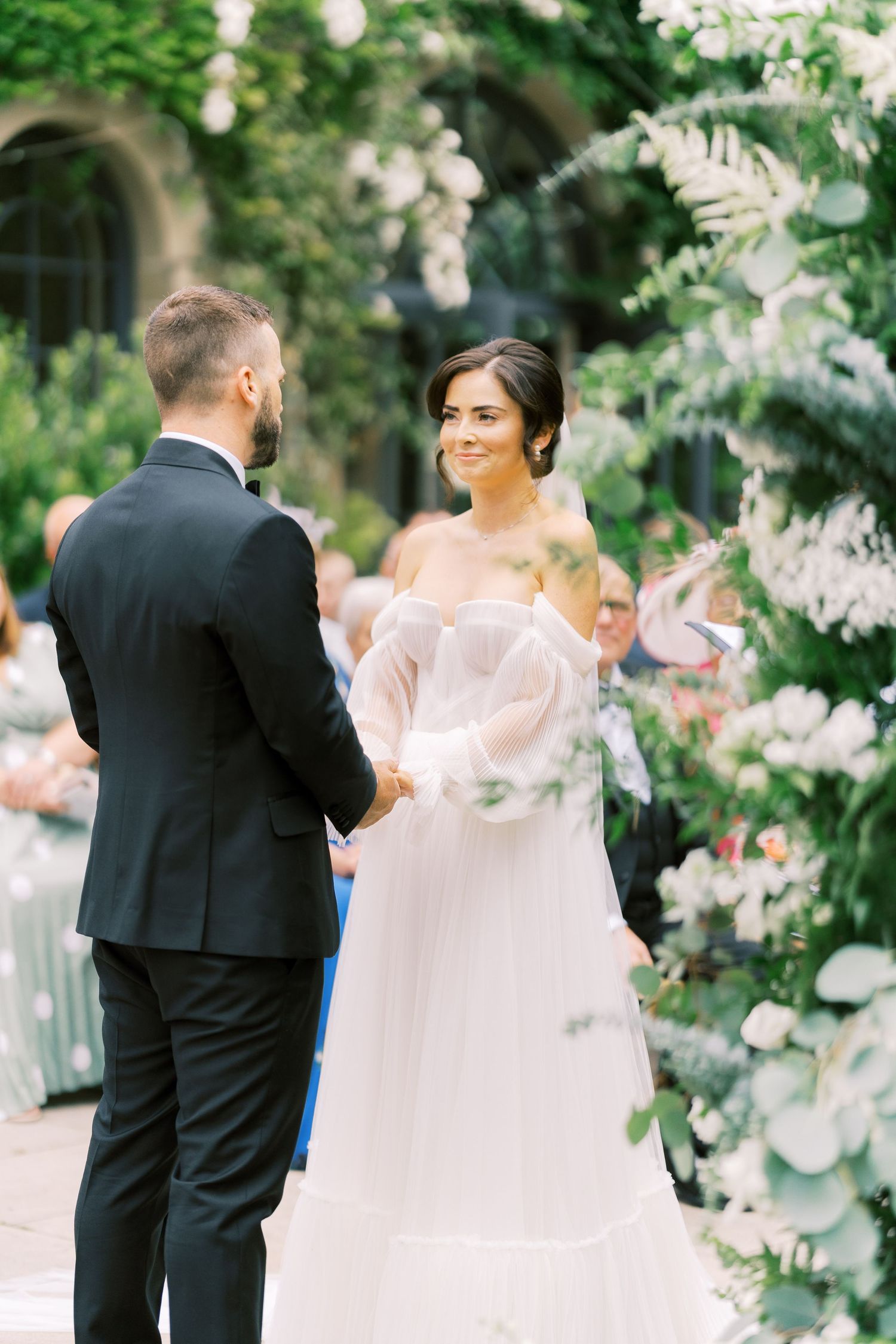 Bride in off-shoulder white gown smiles at groom in black tuxedo during outdoor wedding ceremony with floral arch.