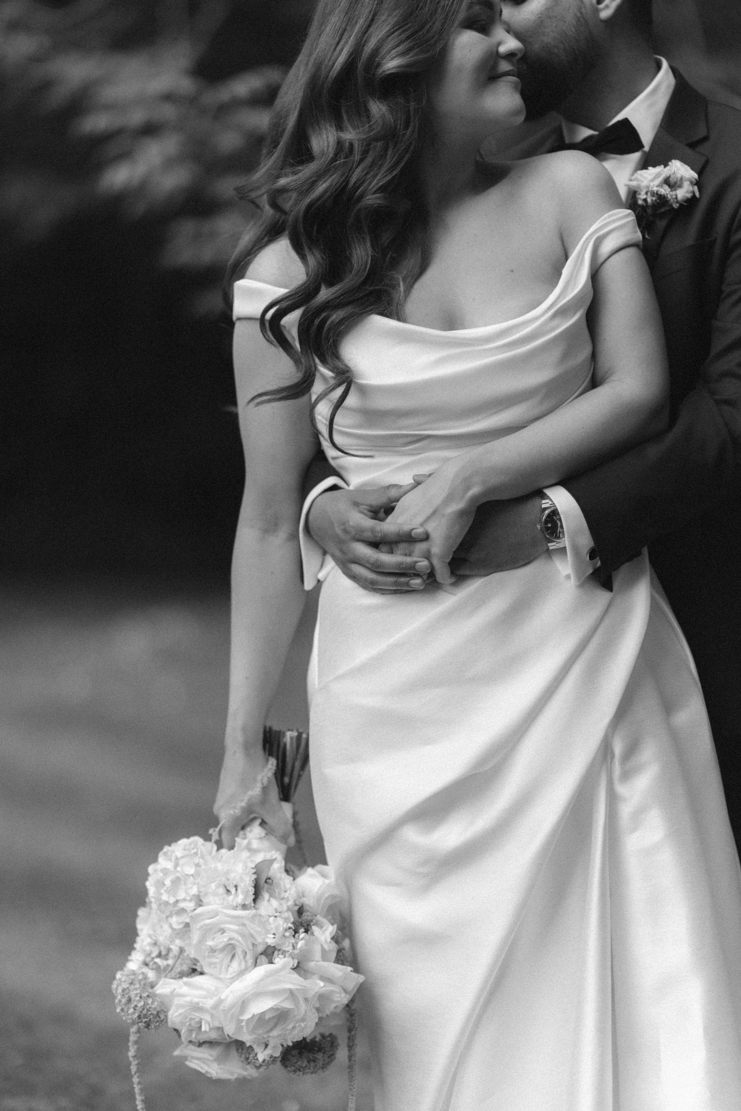 Black and white photo of bride in off-shoulder gown holding bouquet, embraced by groom in suit on wedding day.
