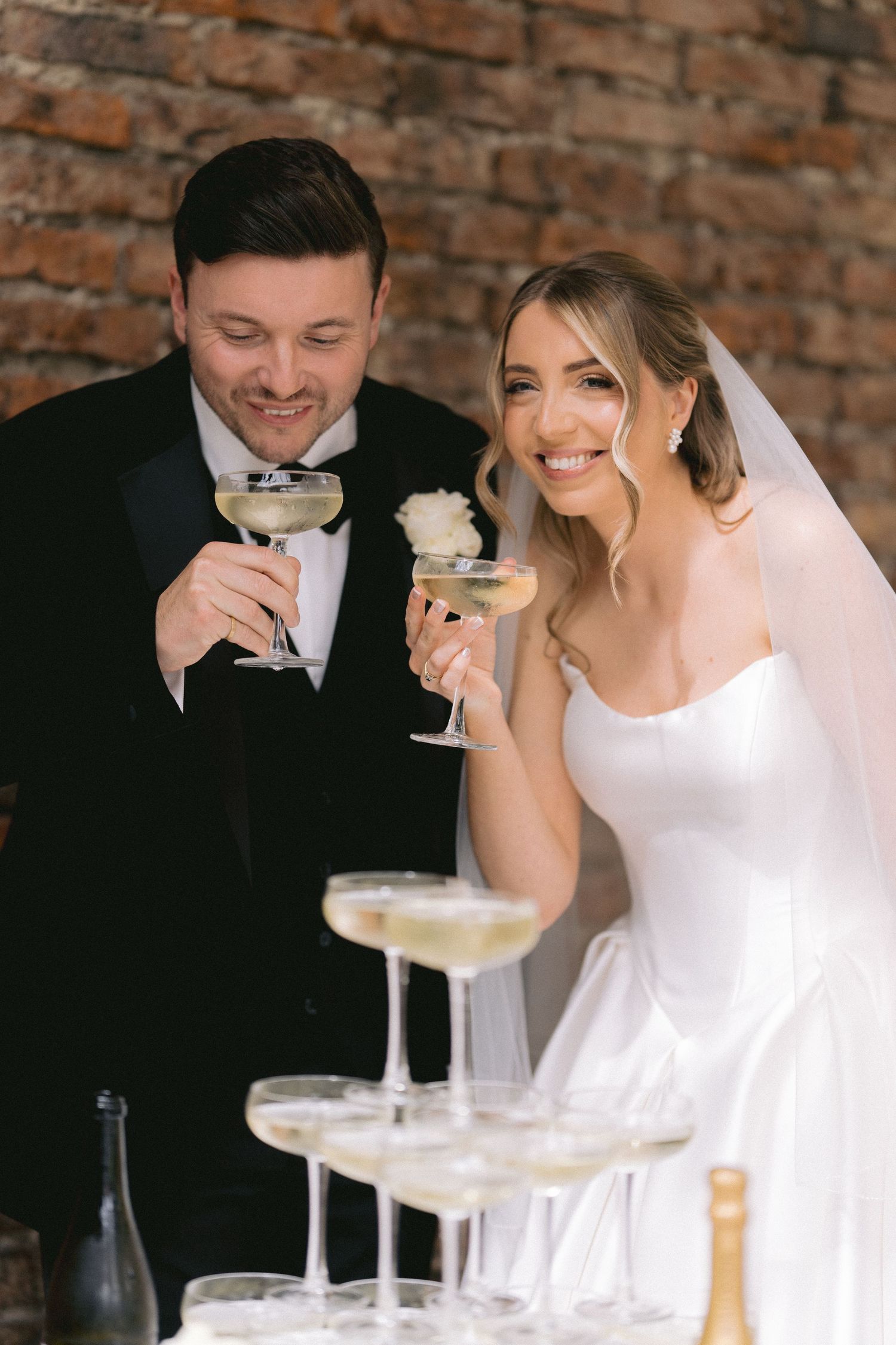Bride and groom smiling while toasting champagne coupe glasses beside a pyramid tower at their wedding reception.
