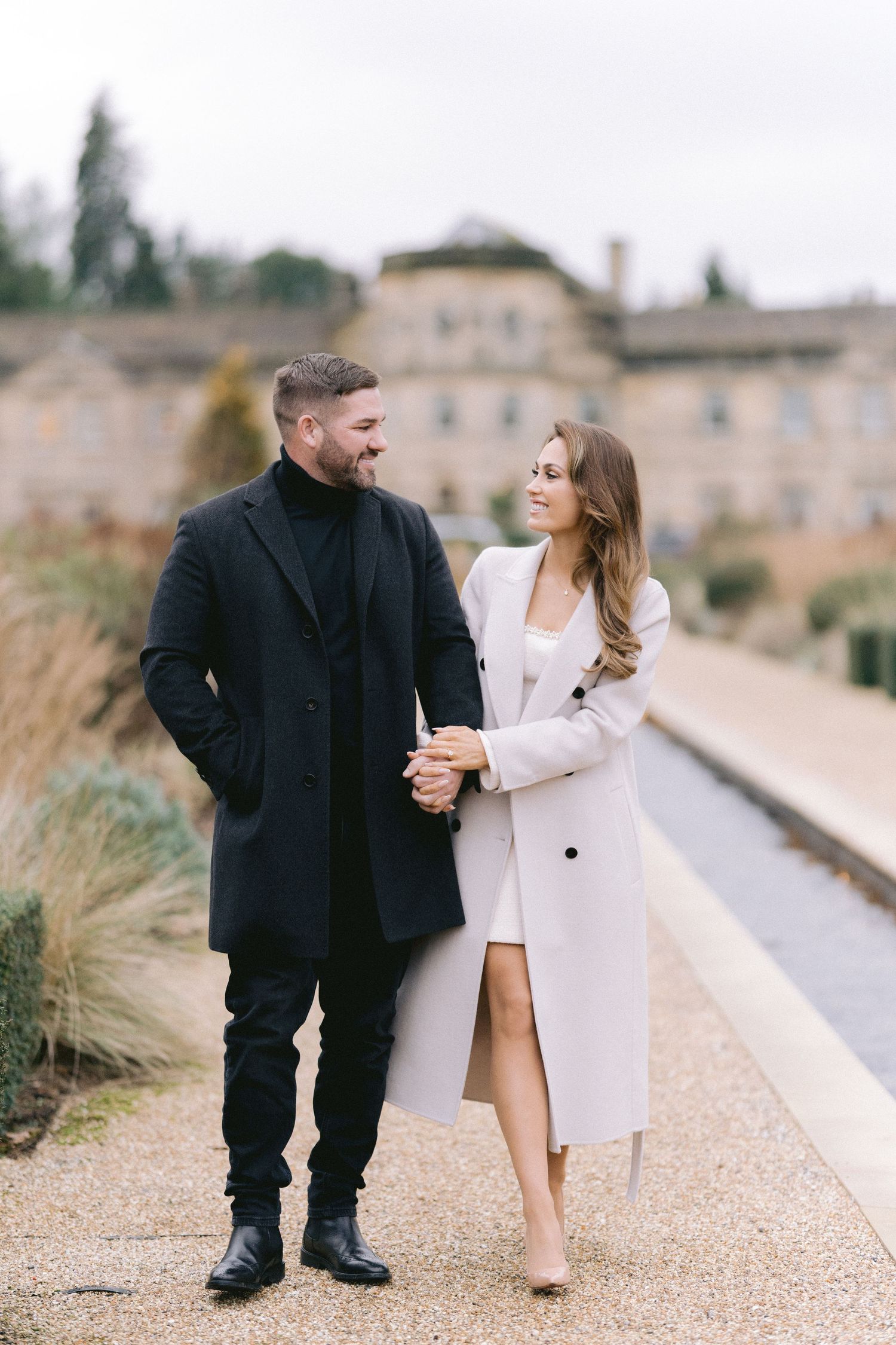 Stylish couple walks hand in hand along garden path, smiling at each other in autumn setting.
