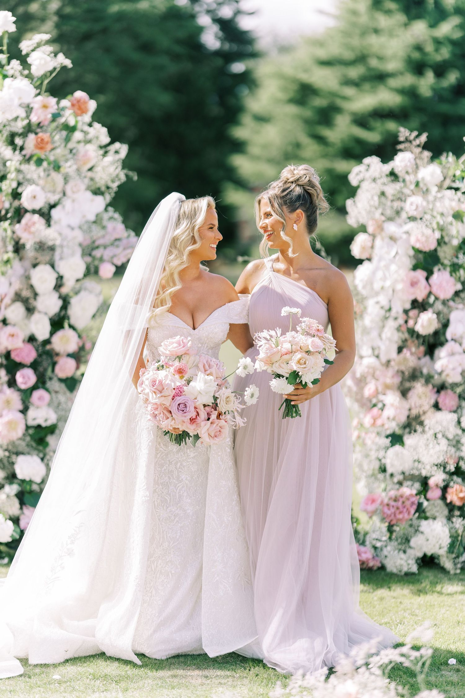 Bride in white gown and bridesmaid in pink dress holding floral bouquets before a floral arch outdoors.