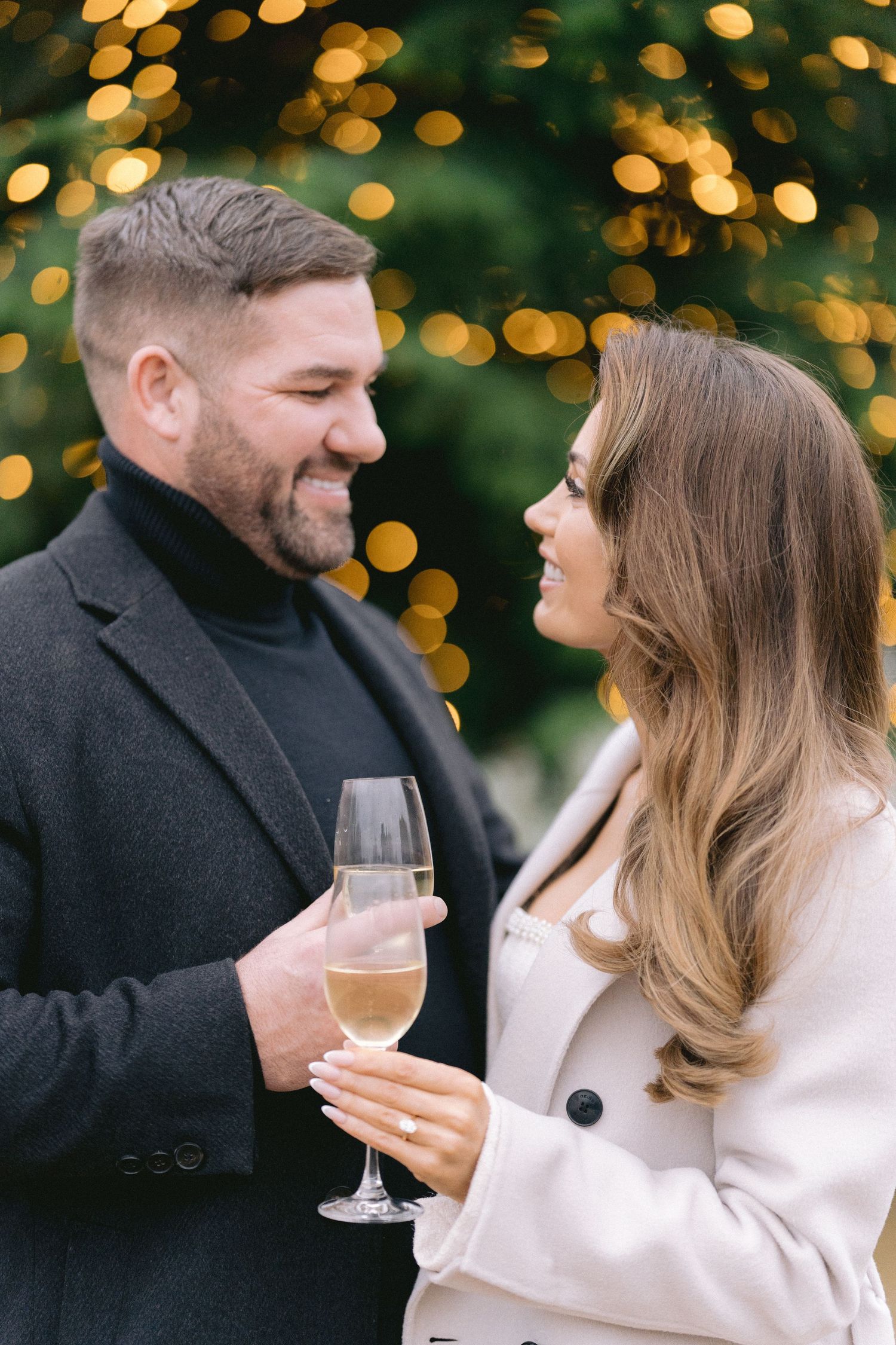 Happy couple sharing a glass of champagne while smiling at each other against a glowing Christmas tree backdrop.