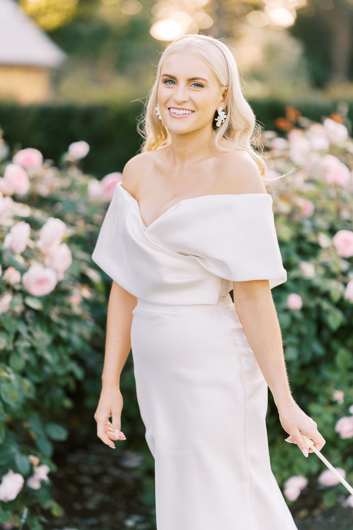 Smiling bride in elegant off-shoulder white gown standing among pink roses in a garden.
