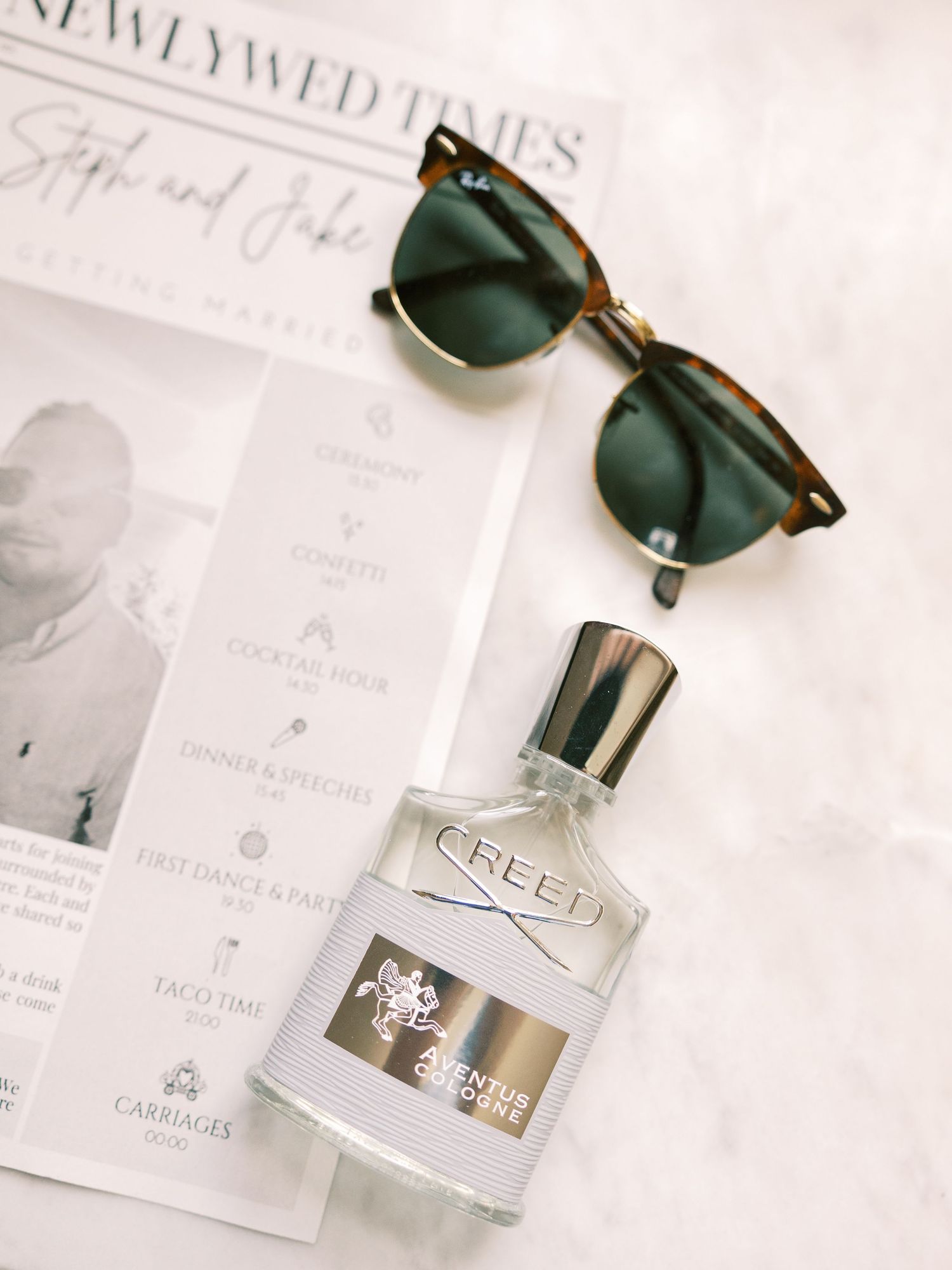 Creed cologne bottle beside Ray-Ban sunglasses on a Newlywed Times wedding program flatlay.