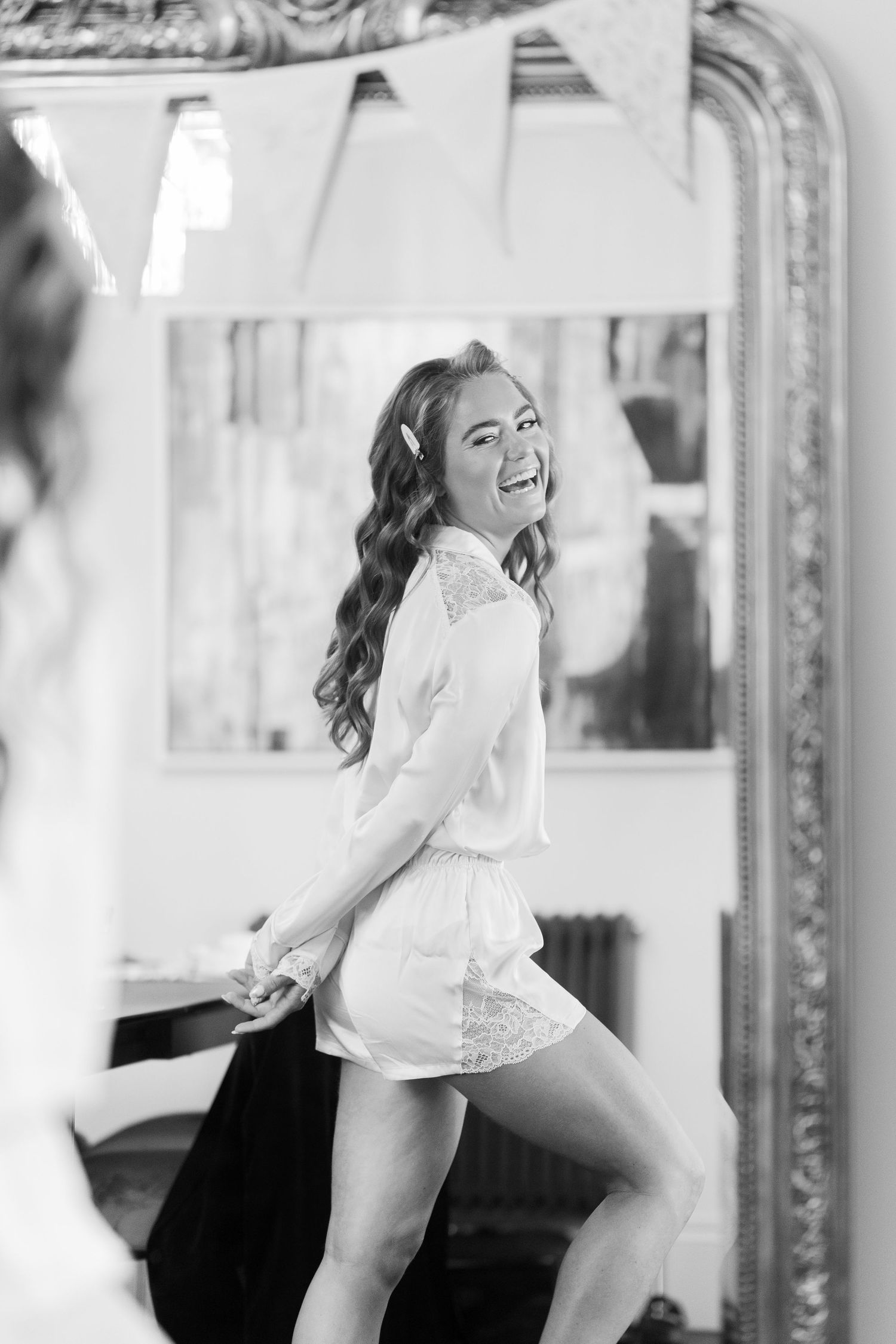 Bride laughing in white lace robe posing in front of ornate mirror in black and white photo.