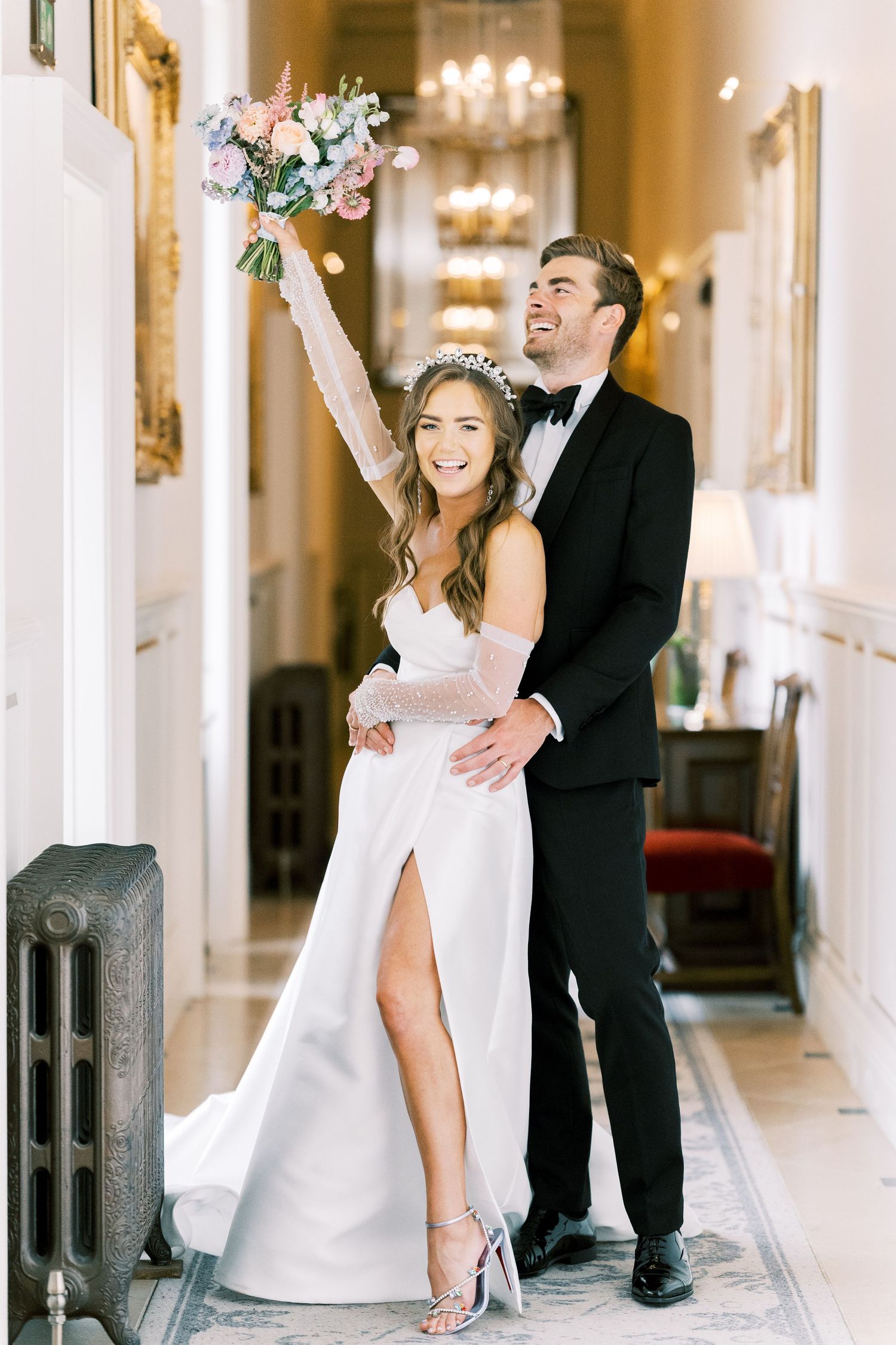 Joyful bride in white gown raises bouquet while groom in tuxedo embraces her in elegant hallway.