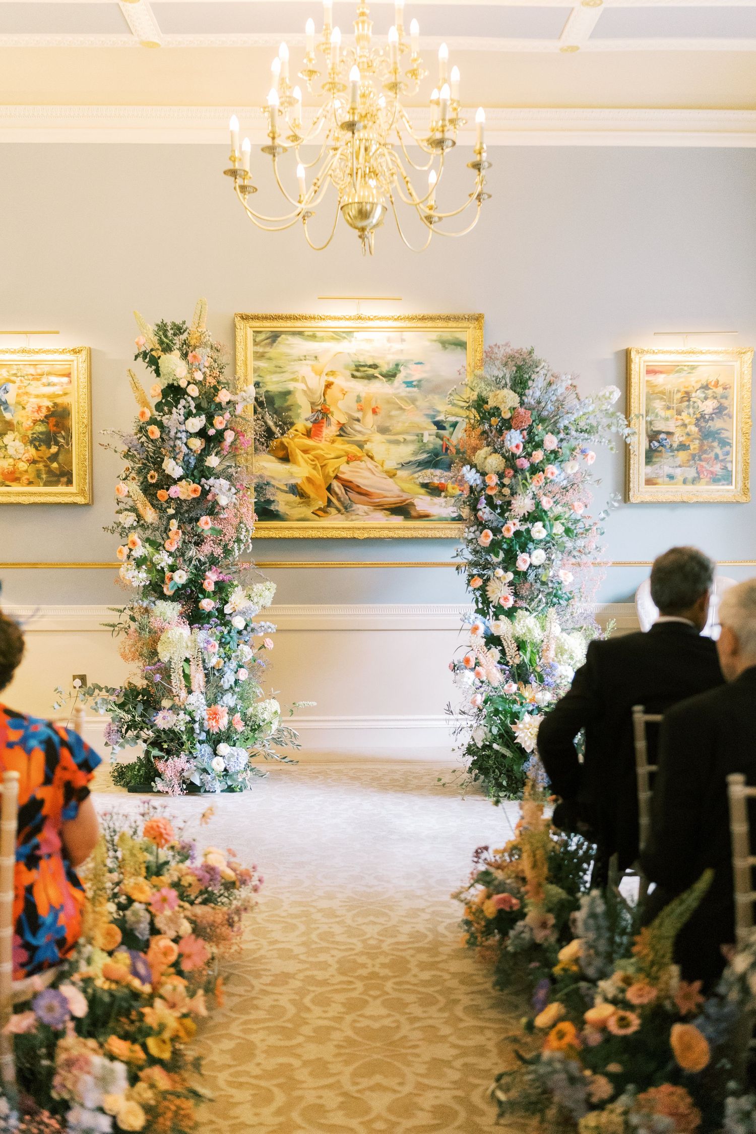 Elegant indoor wedding ceremony with floral arch, aisle flowers, chandelier, and gold-framed paintings in a classic venue.