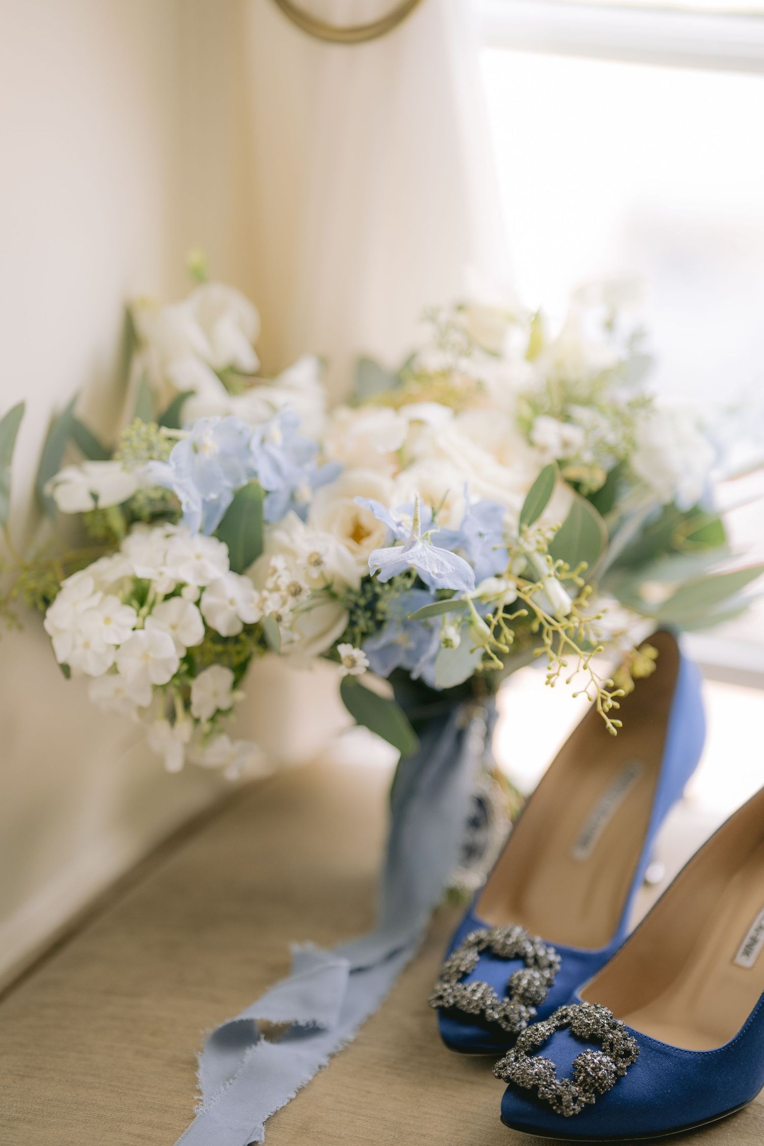 Blue satin Manolo Blahnik heels with crystal buckles beside a white and blue hydrangea bridal bouquet with silk ribbon.