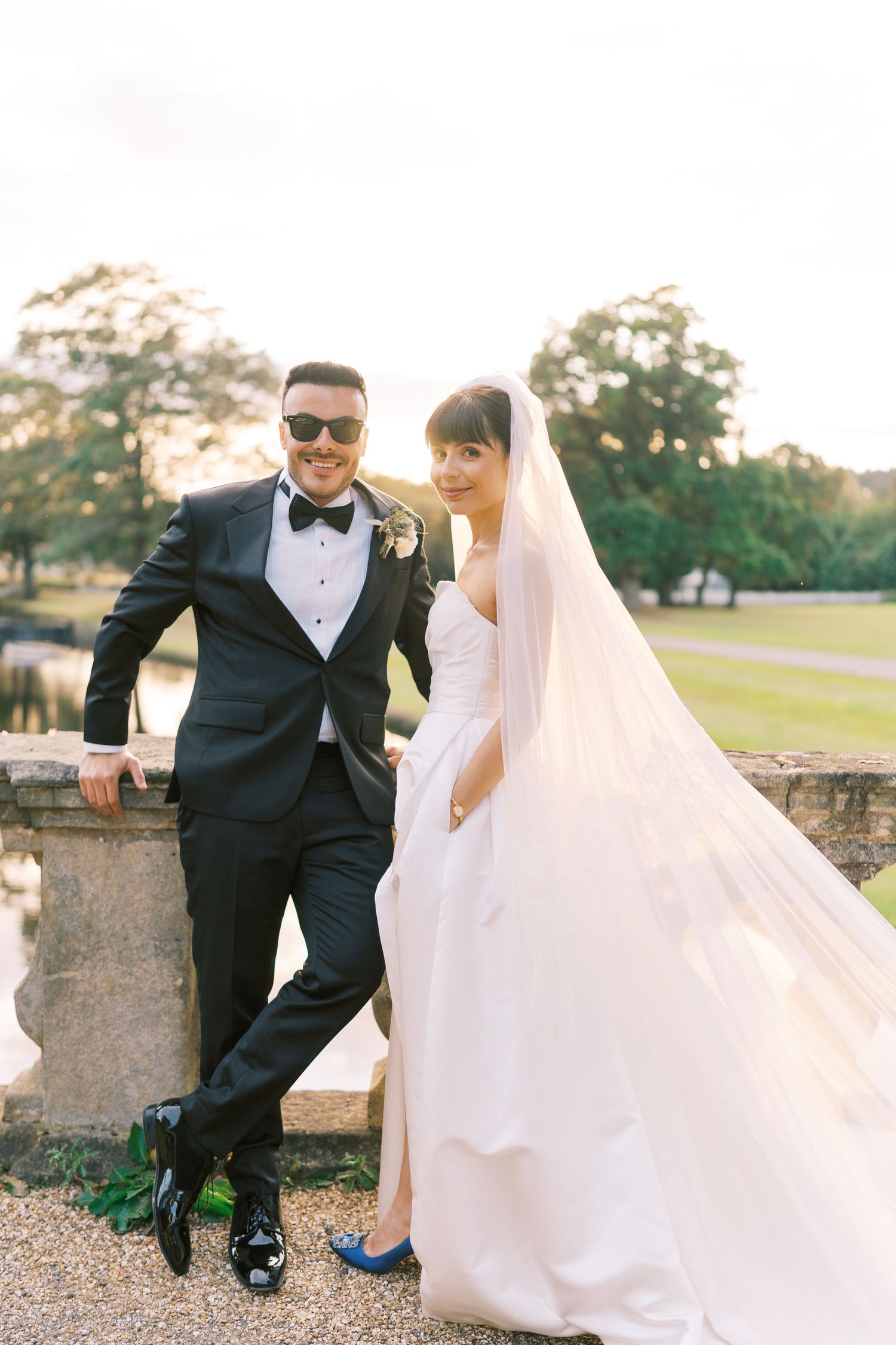 Elegant bride and groom posing by stone balustrade, he in tuxedo with sunglasses, she in white gown with veil.