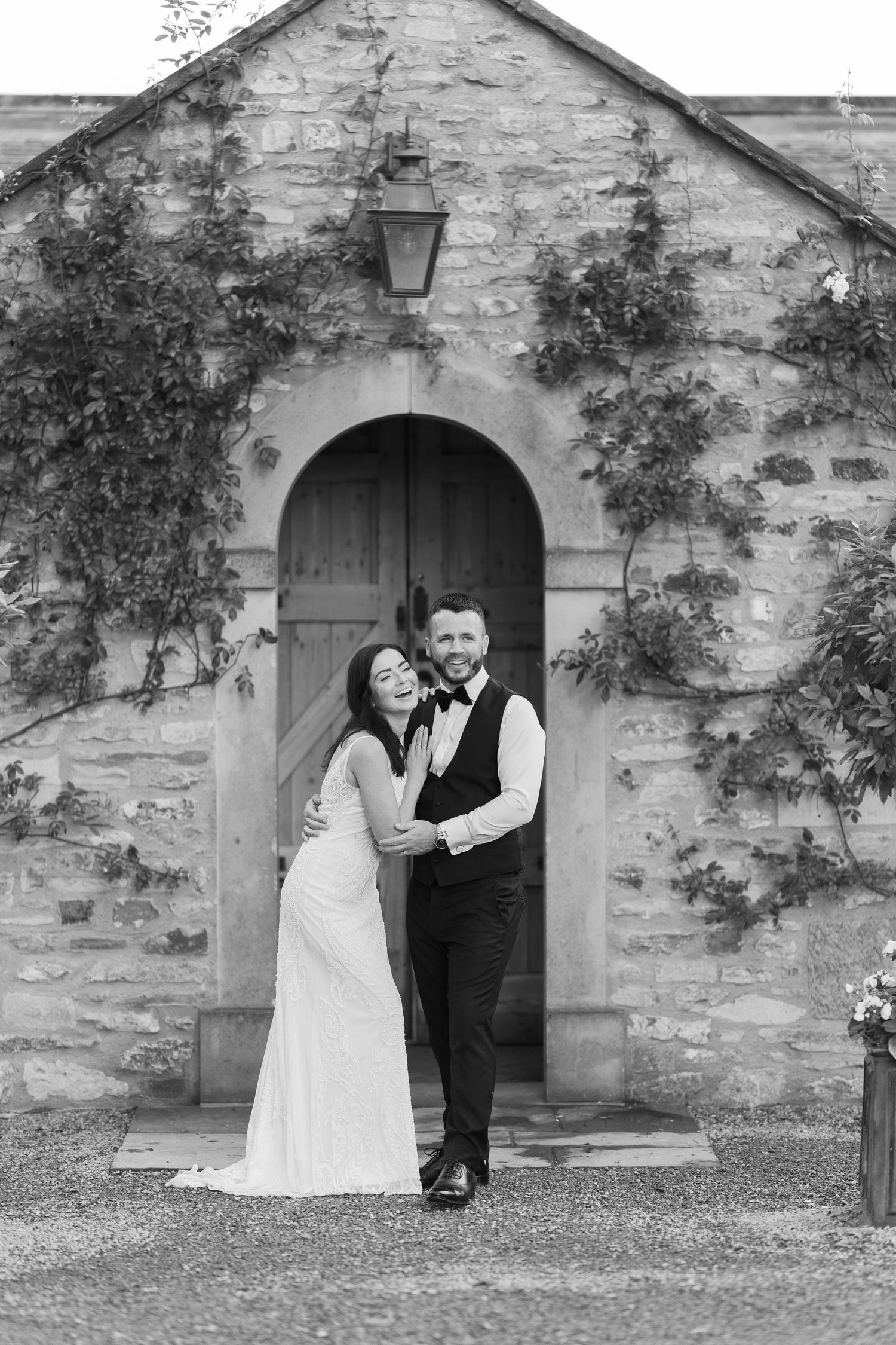 Black and white photo of bride and groom embracing before a stone arch doorway with ivy walls.