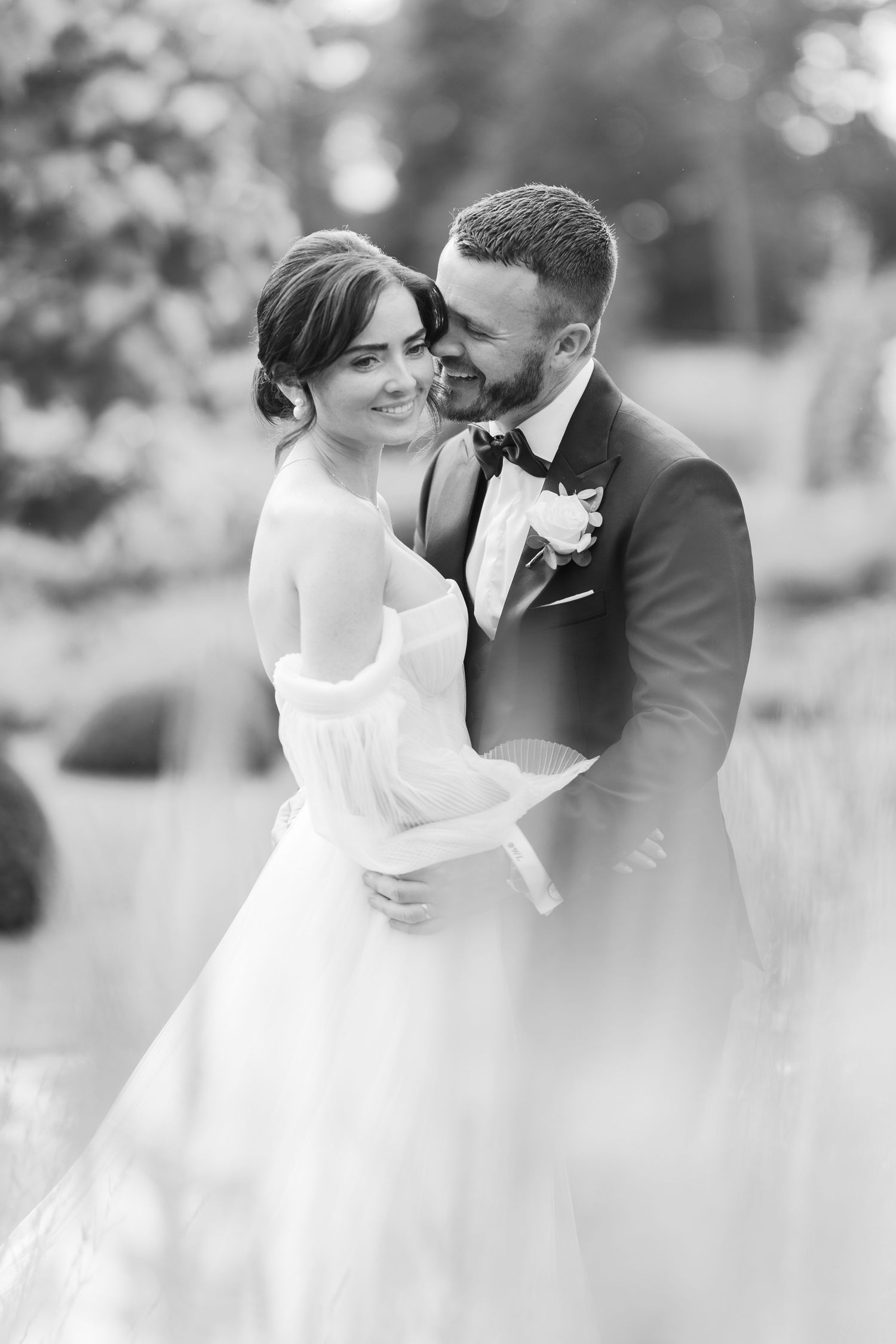 Black and white photo of smiling bride in off-shoulder gown embraced by groom in tuxedo outdoors.