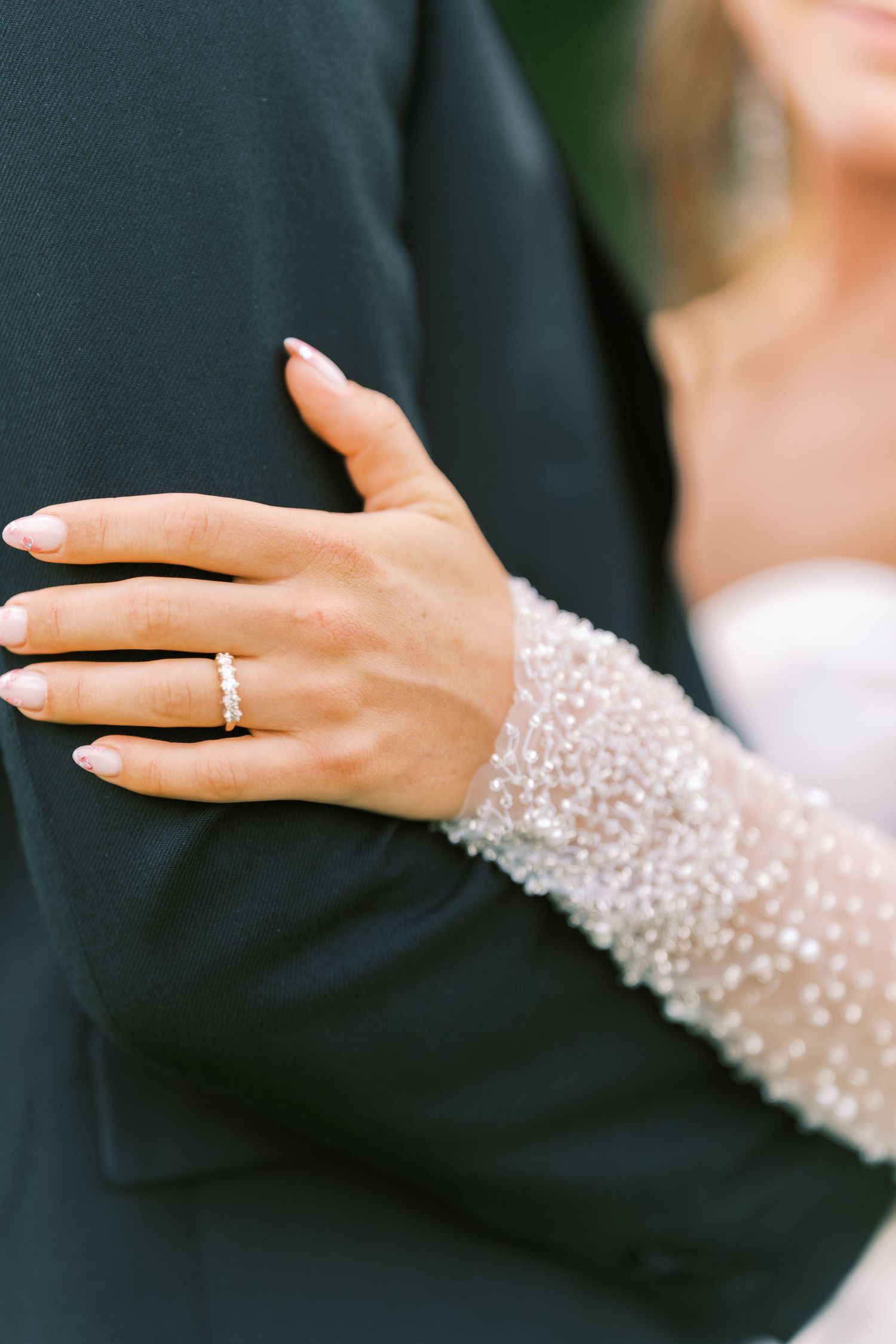 Close-up of bride's hand wearing gold wedding band resting on groom's dark suit jacket sleeve.