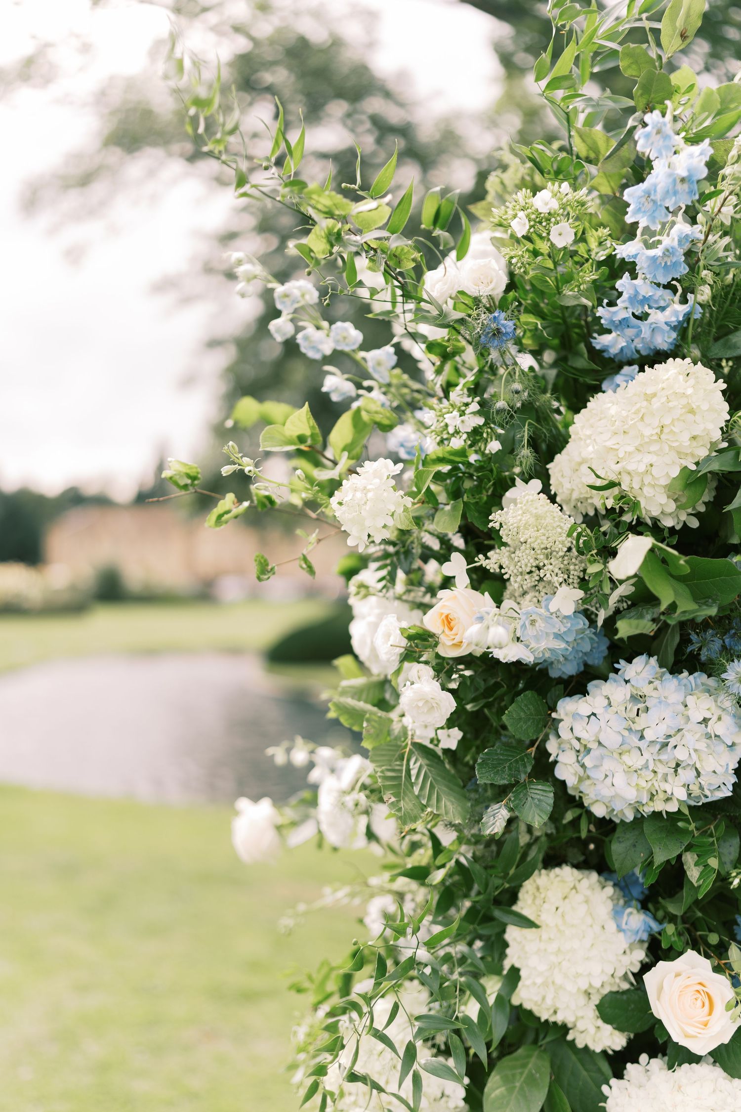 Close-up of wedding arch flowers with white hydrangeas, blue delphinium, and peach roses in a garden setting.