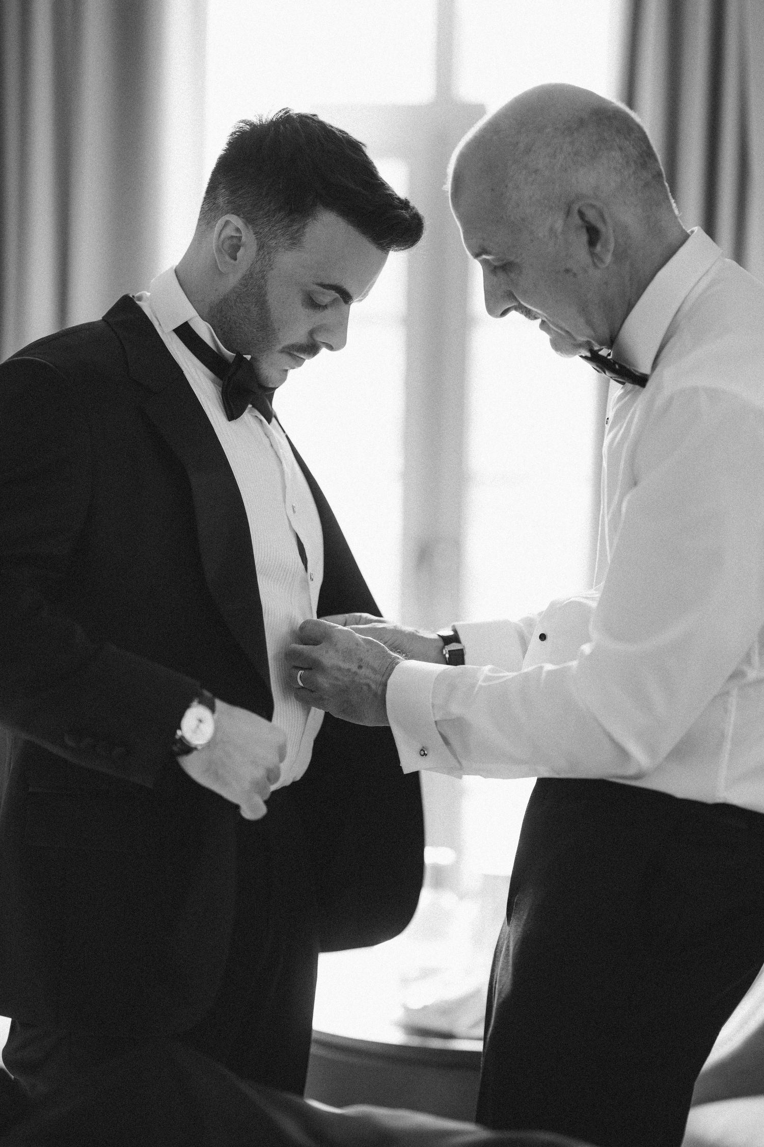 Black and white photo of groom in tuxedo being helped by older man in bow tie during wedding preparations.