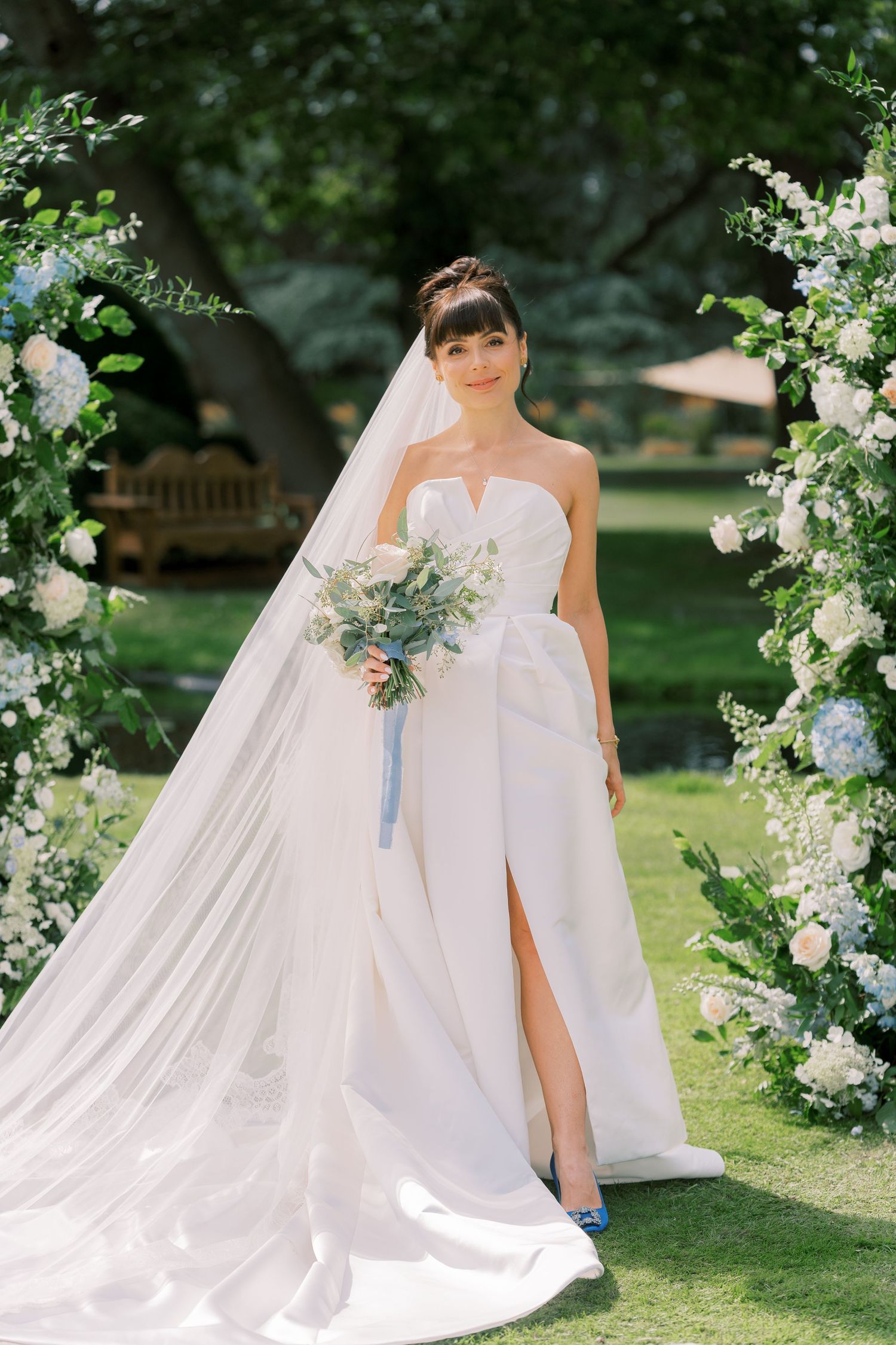 Bride in strapless white gown with slit and veil holds bouquet by floral arch outdoors.