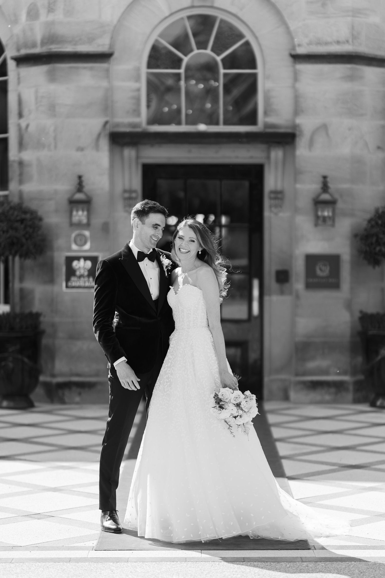 Black and white photo of a bride and groom posing elegantly outside a grand stone building on their wedding day.