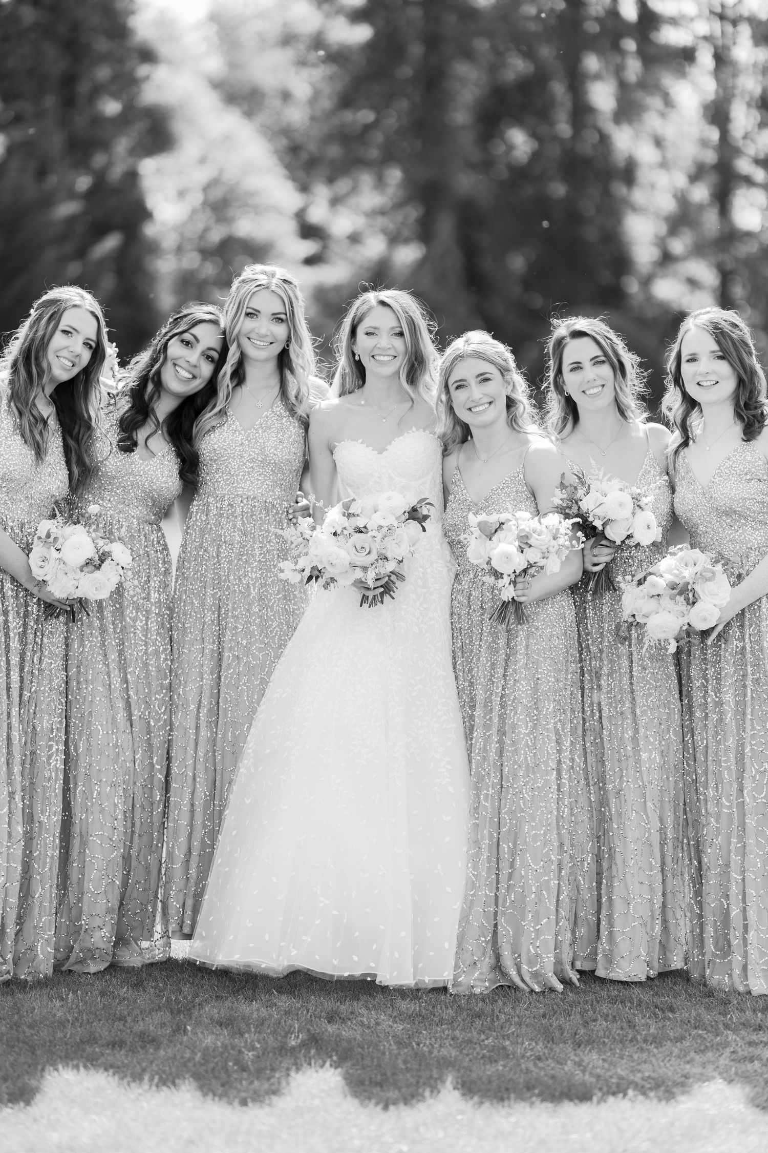 Black and white photo of bride in ballgown with six bridesmaids in sequin dresses holding bouquets outside.