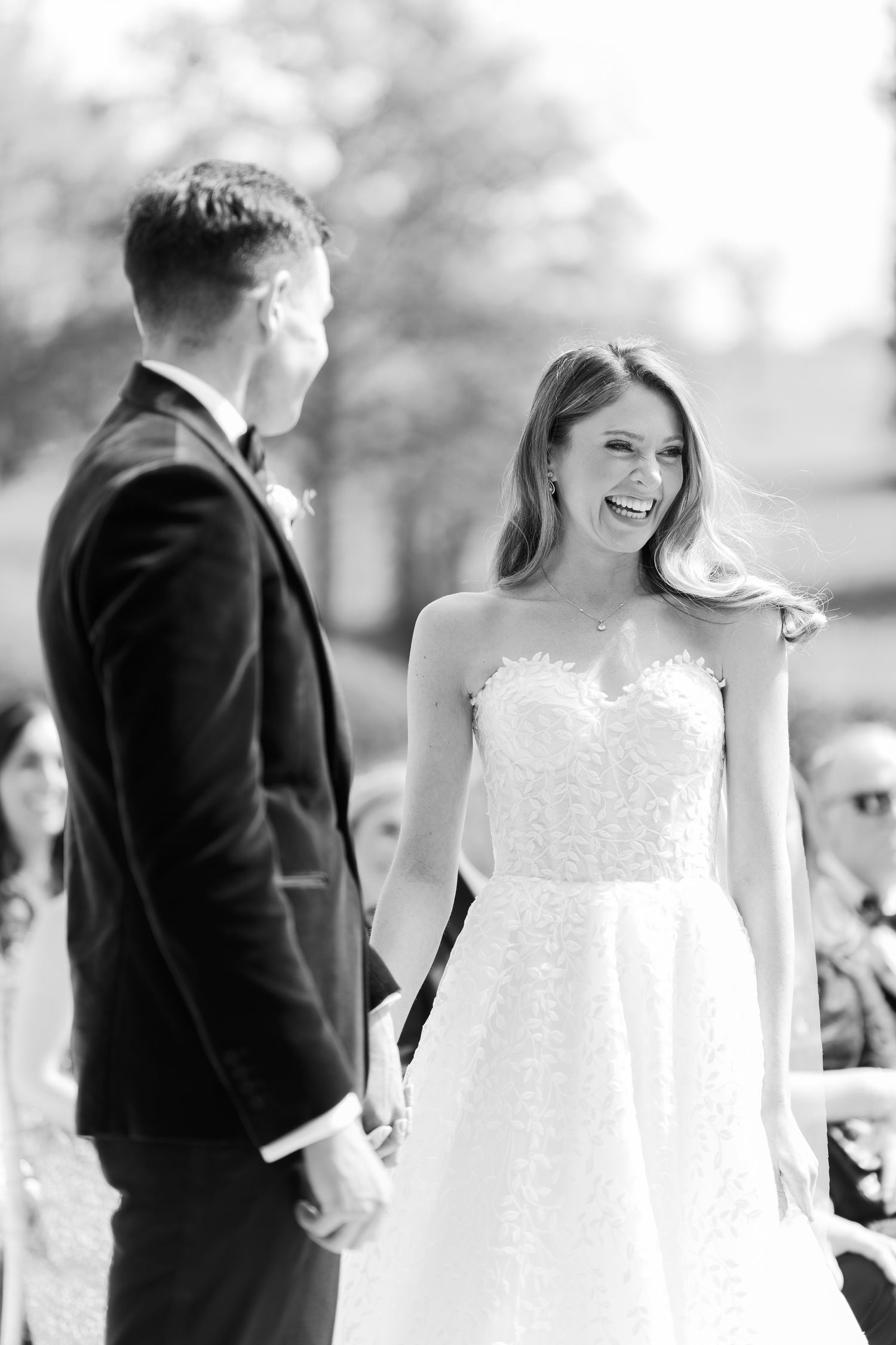 Black and white photo of a joyful bride laughing while holding hands with groom during outdoor wedding ceremony.