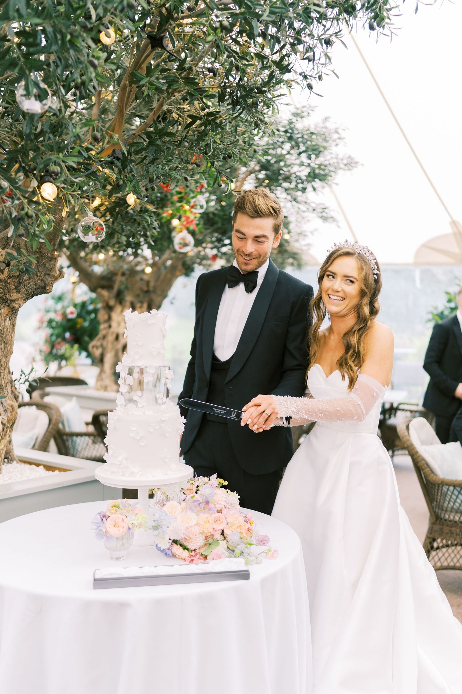 Bride and groom cutting white tiered wedding cake under olive tree at elegant outdoor reception.