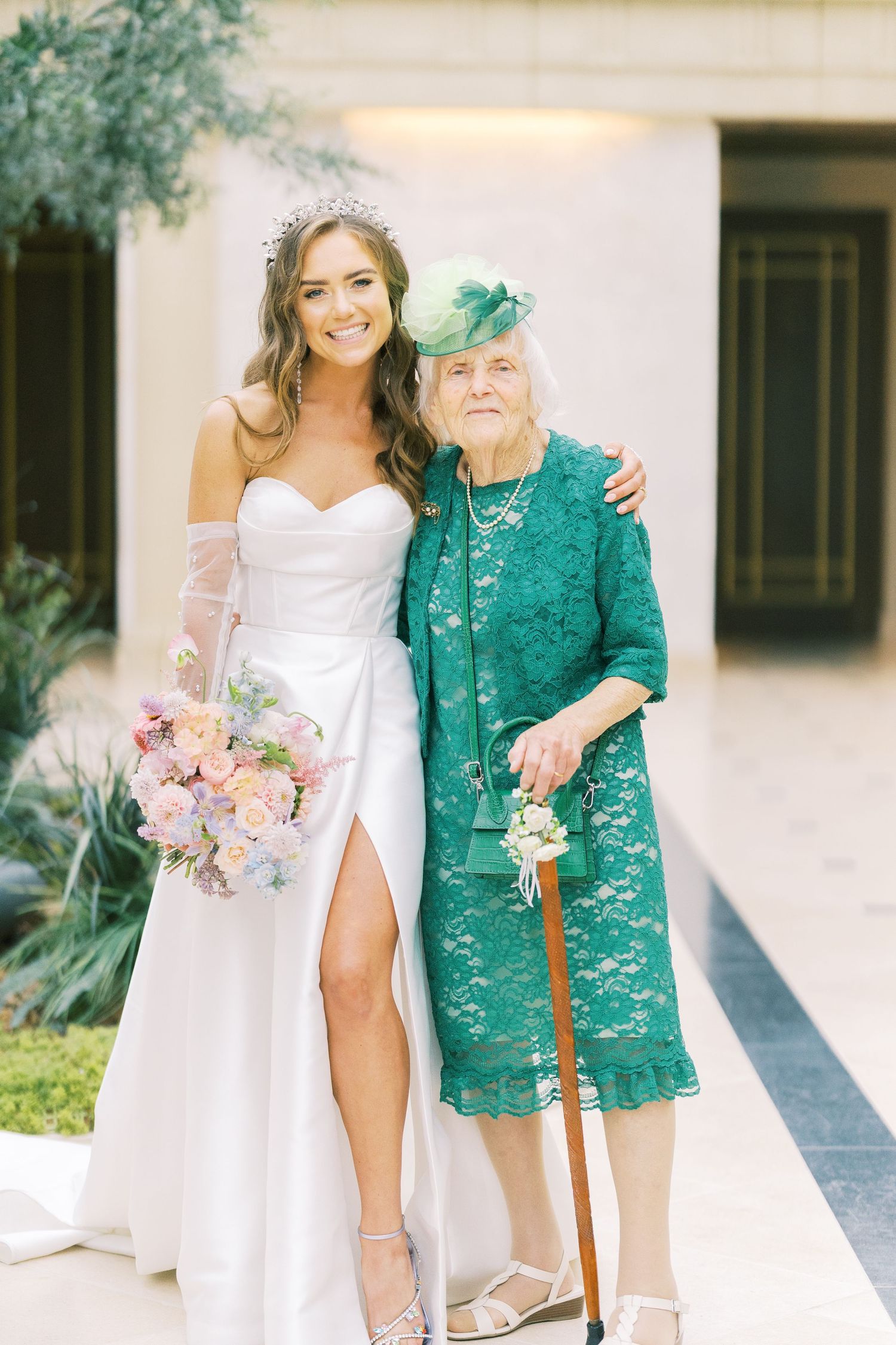Bride in white gown with pink bouquet poses with elderly woman in green lace dress and hat at wedding.