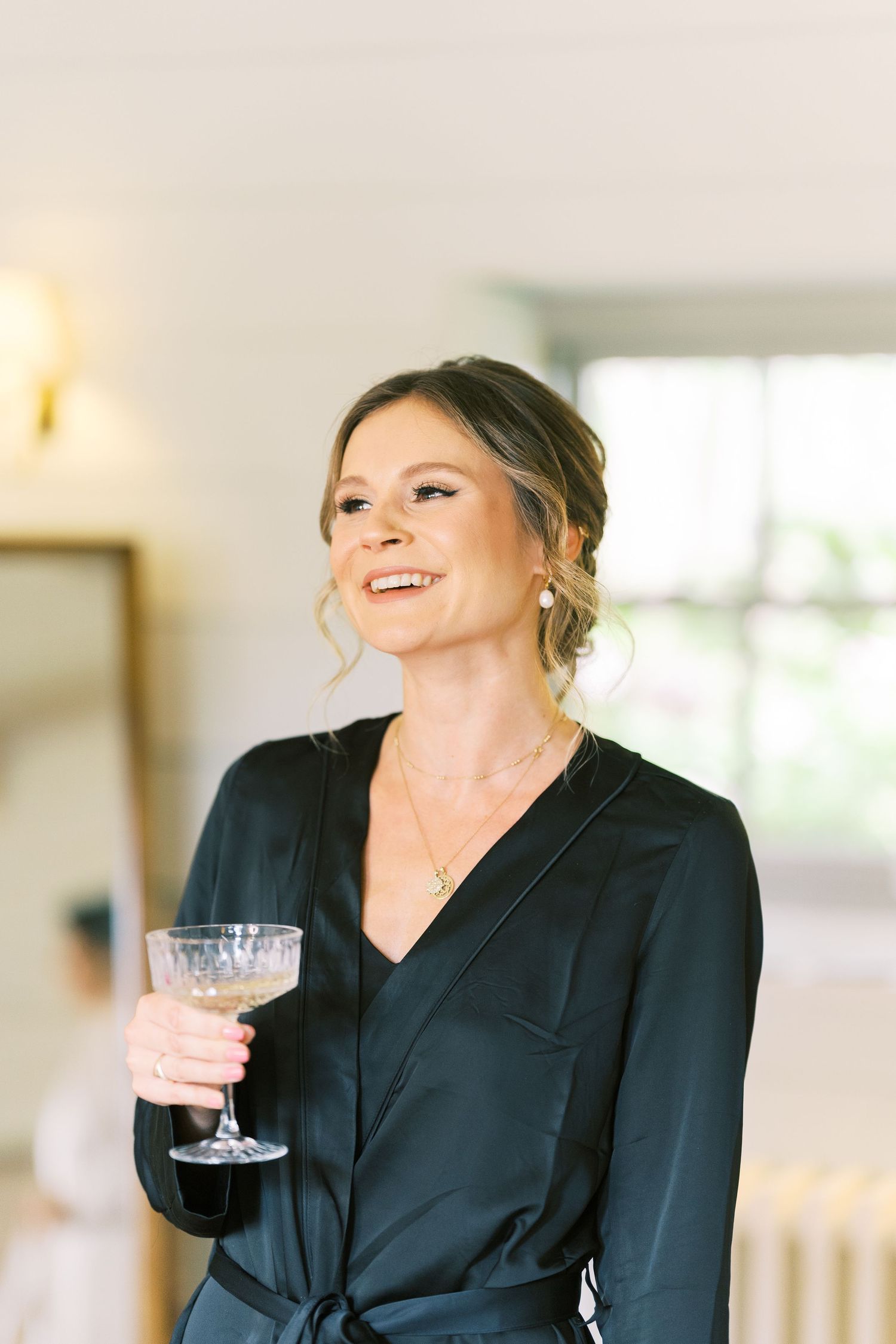 Smiling bridesmaid in black satin robe holds a cocktail glass during wedding preparations.