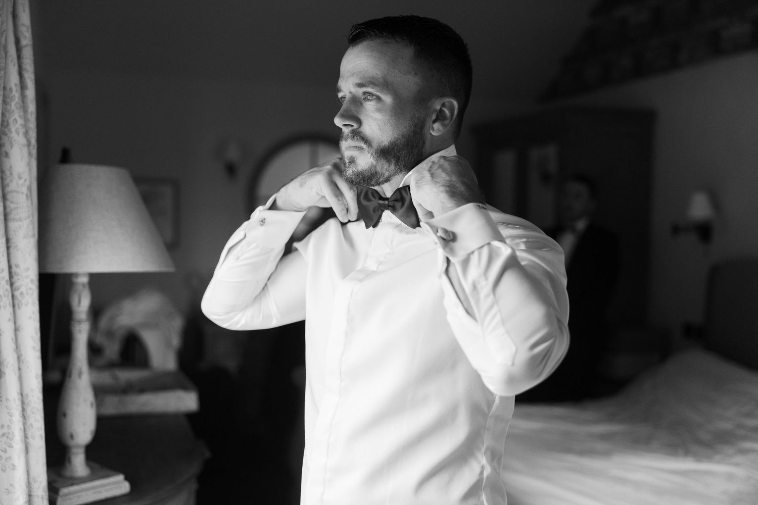 Groom in white dress shirt adjusting bow tie in bedroom mirror, black and white wedding preparation photo.