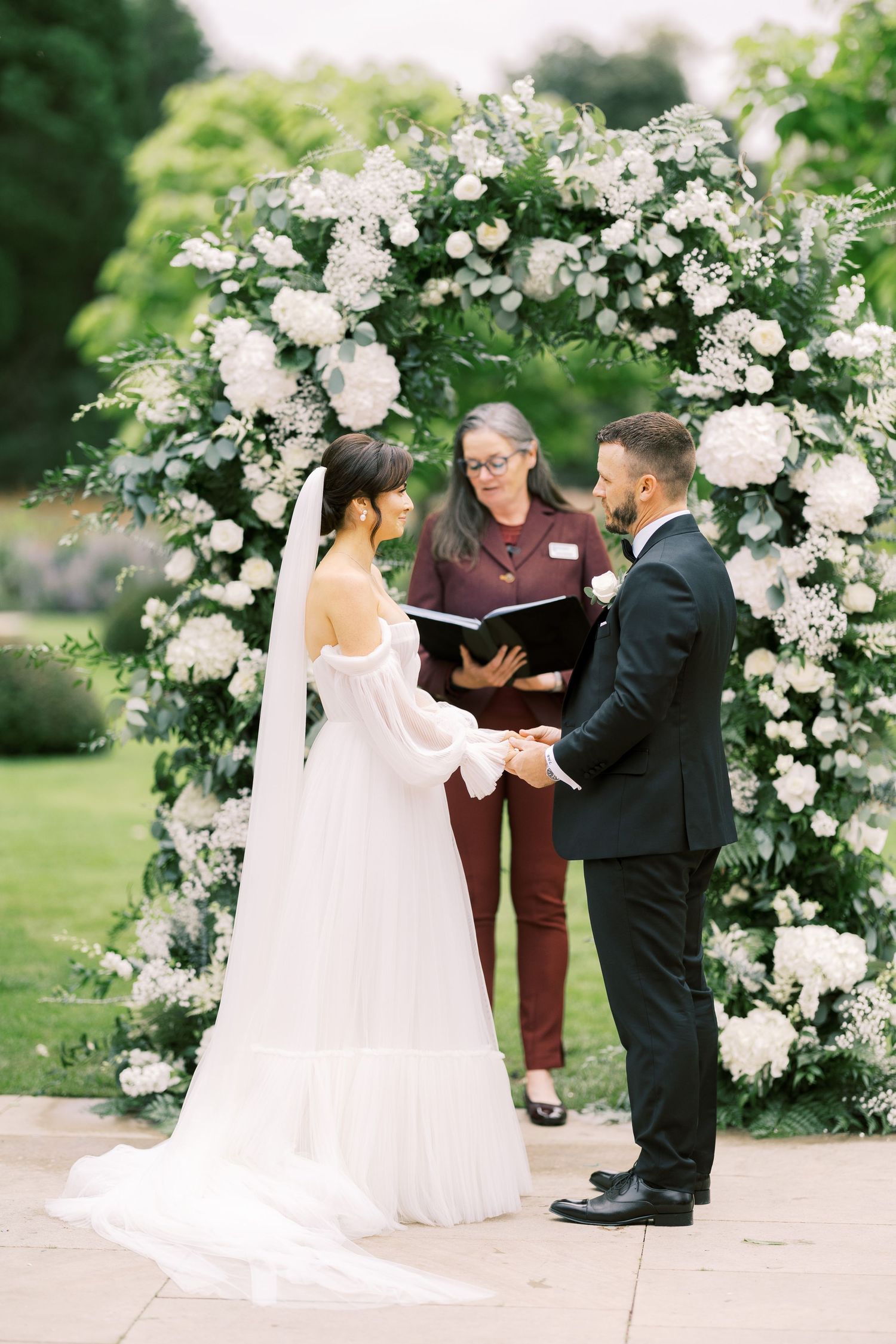 Bride and groom exchange vows under a white floral arch outdoors, officiated by a woman in burgundy.