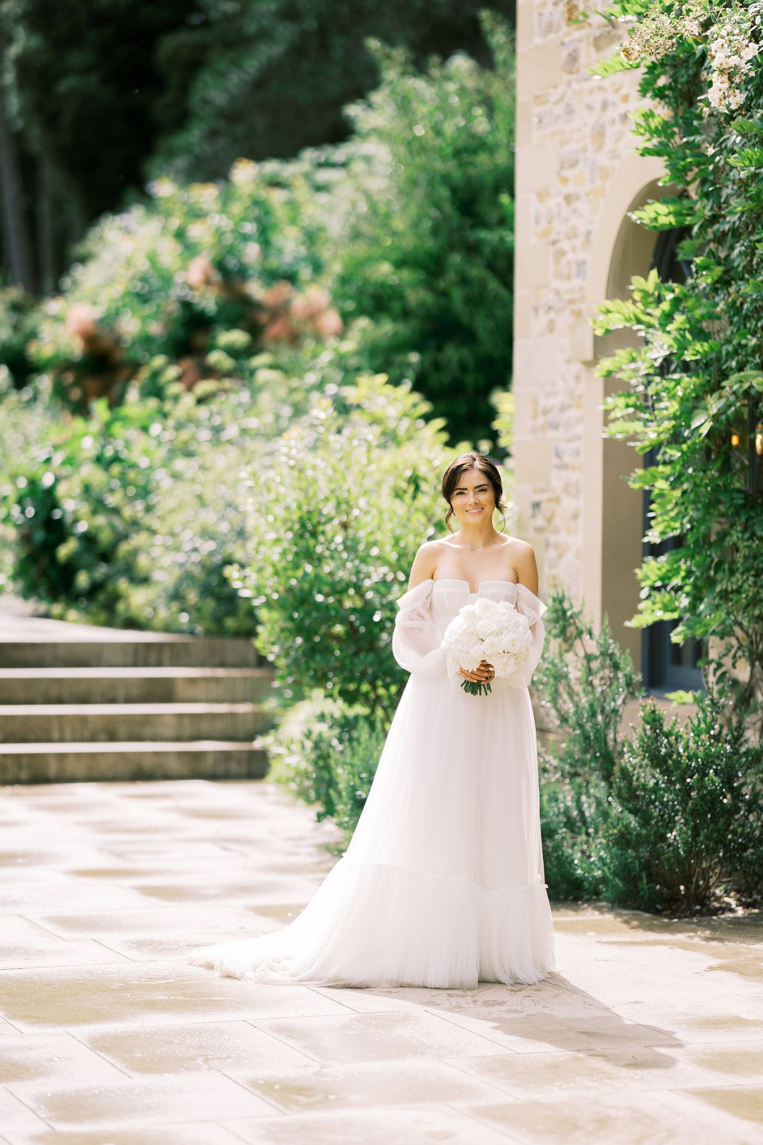 Elegant bride in off-shoulder white gown holding bouquet, standing in lush garden beside stone building.