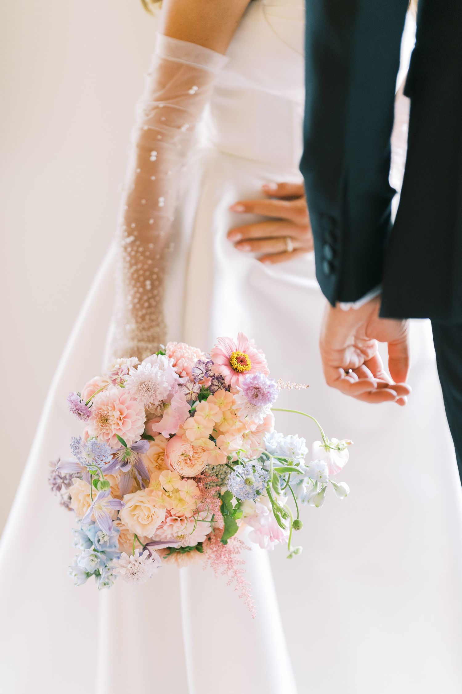 Close-up of bride holding soft pastel wedding bouquet with pink roses and lavender blooms beside groom.