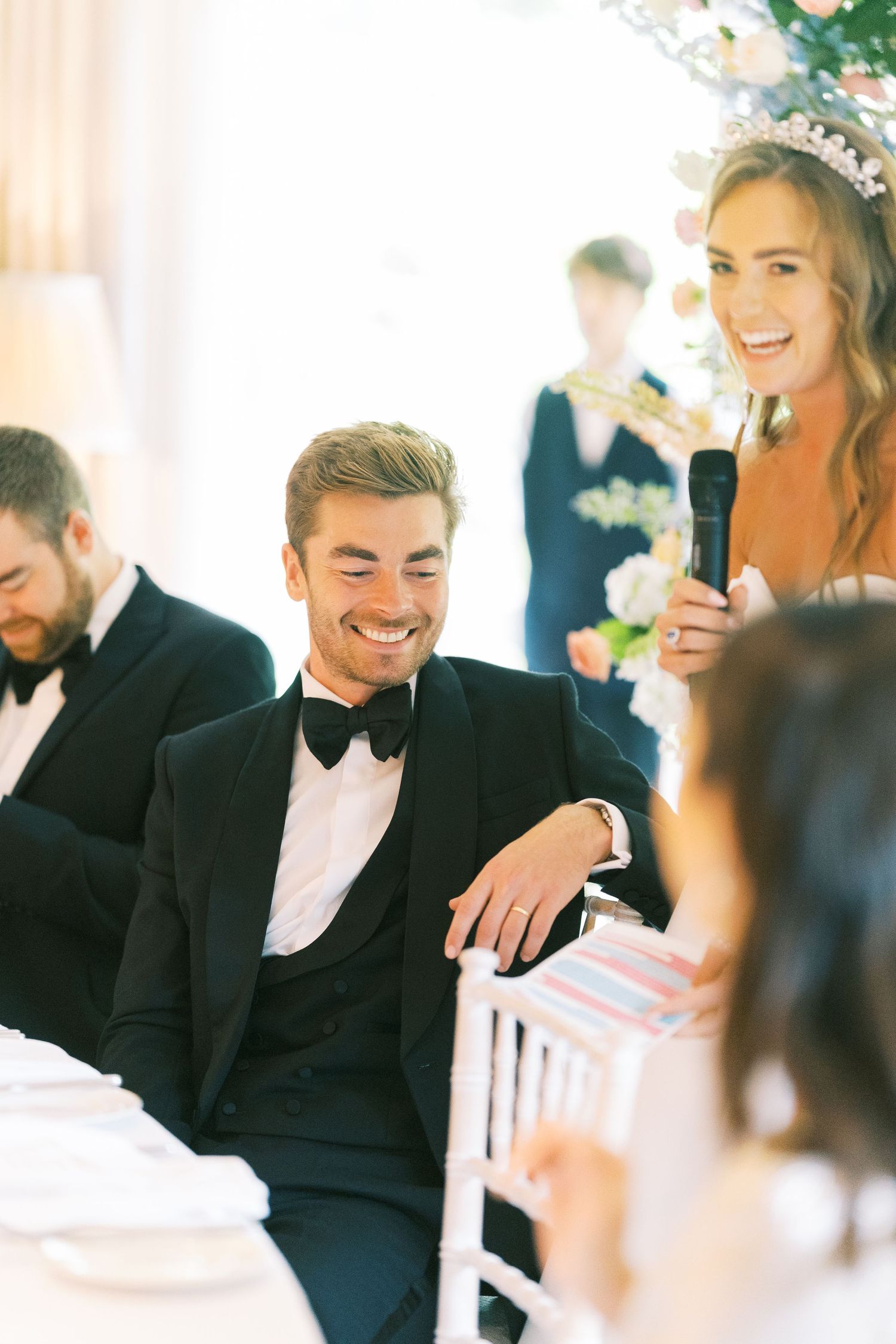 Smiling groom in black tuxedo sits at wedding reception table as bridesmaid holds microphone giving a toast.
