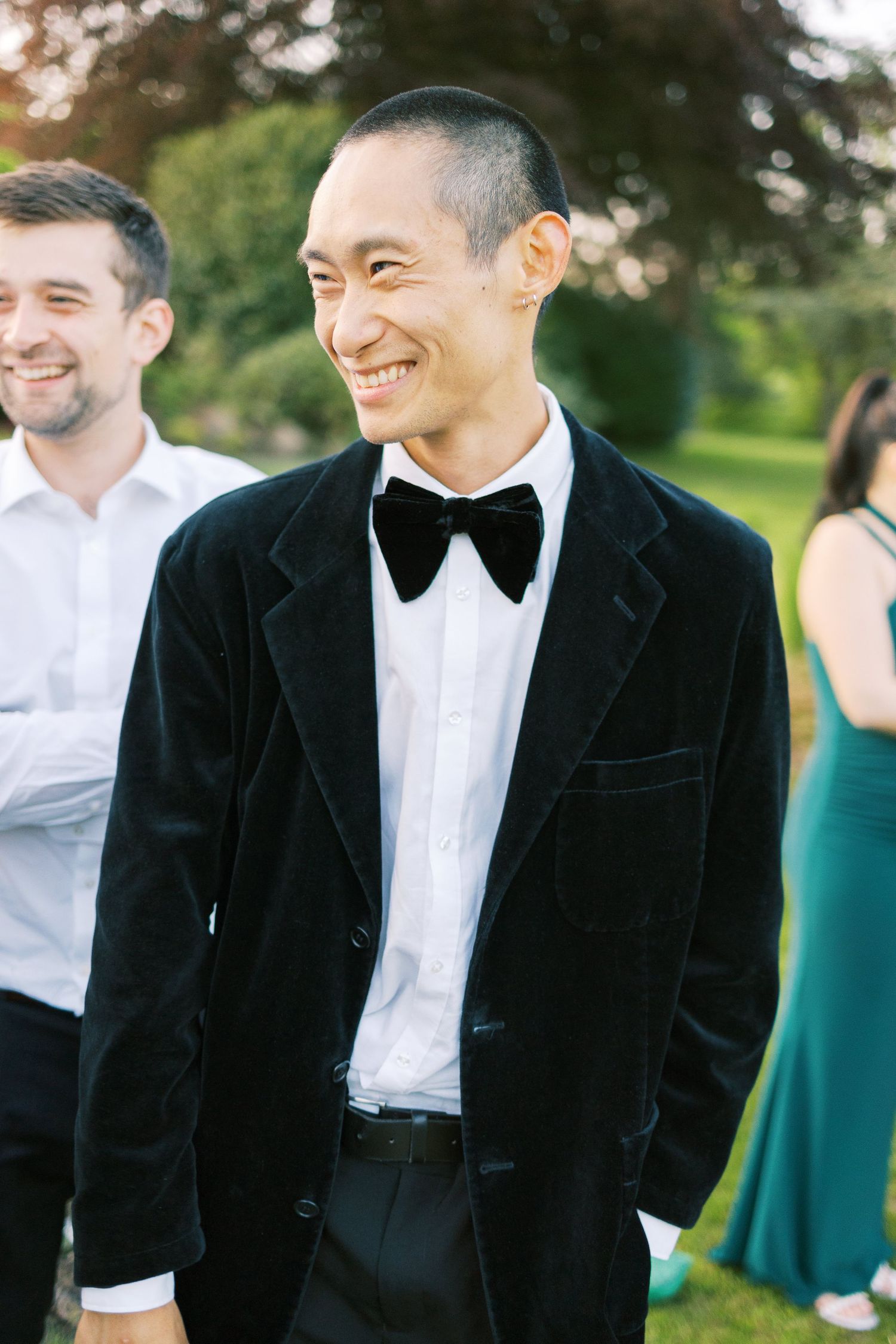 Smiling man in black velvet tuxedo with bow tie enjoying an outdoor wedding celebration.