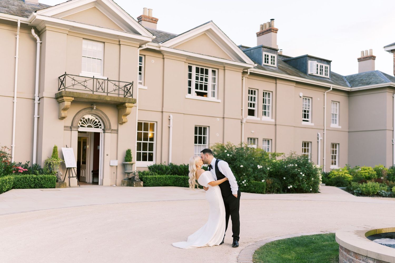 Bride and groom share a romantic dip kiss outside an elegant manor house wedding venue with manicured gardens.