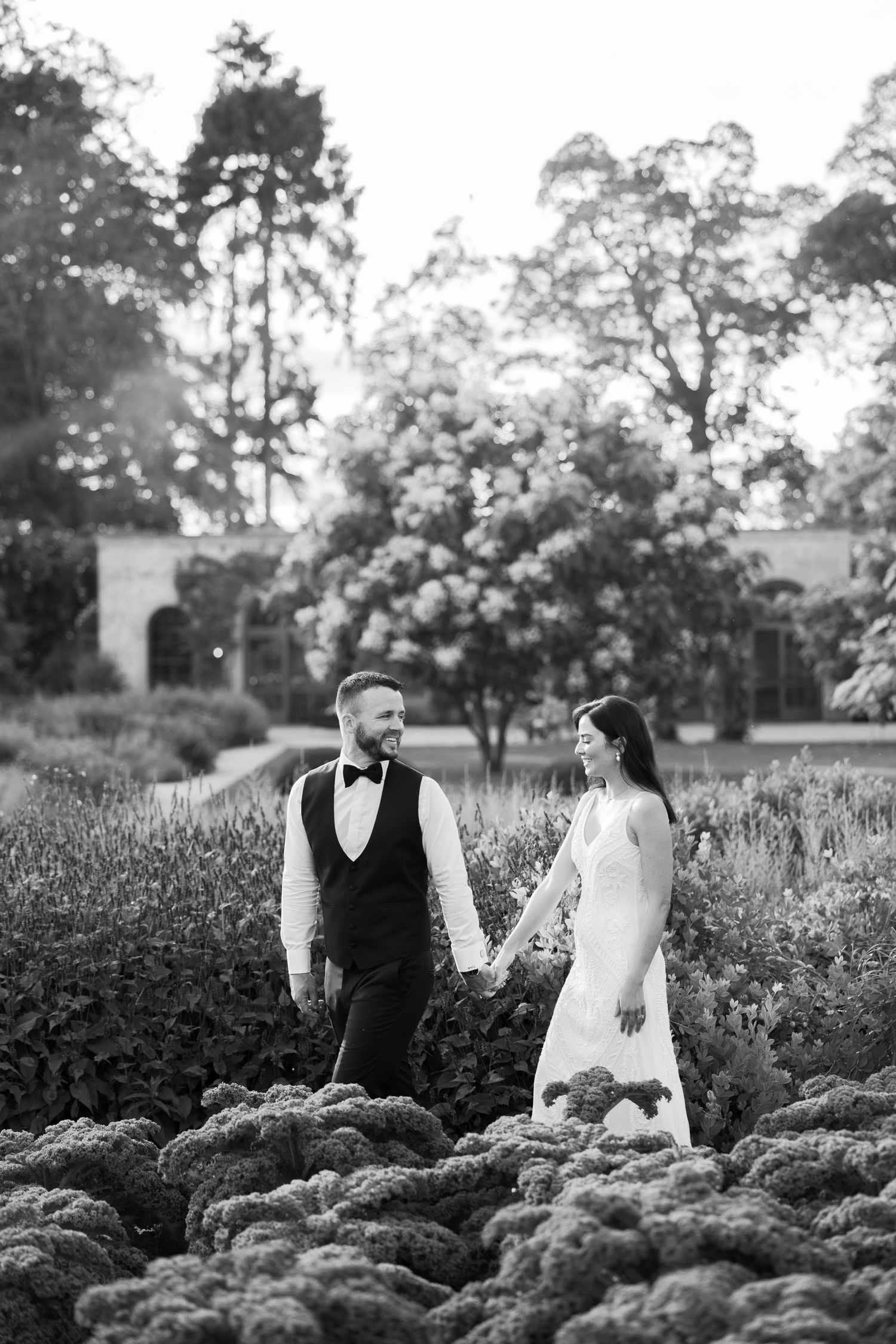 Black and white photo of bride and groom holding hands in a lush garden, surrounded by blooming flowers and trees.