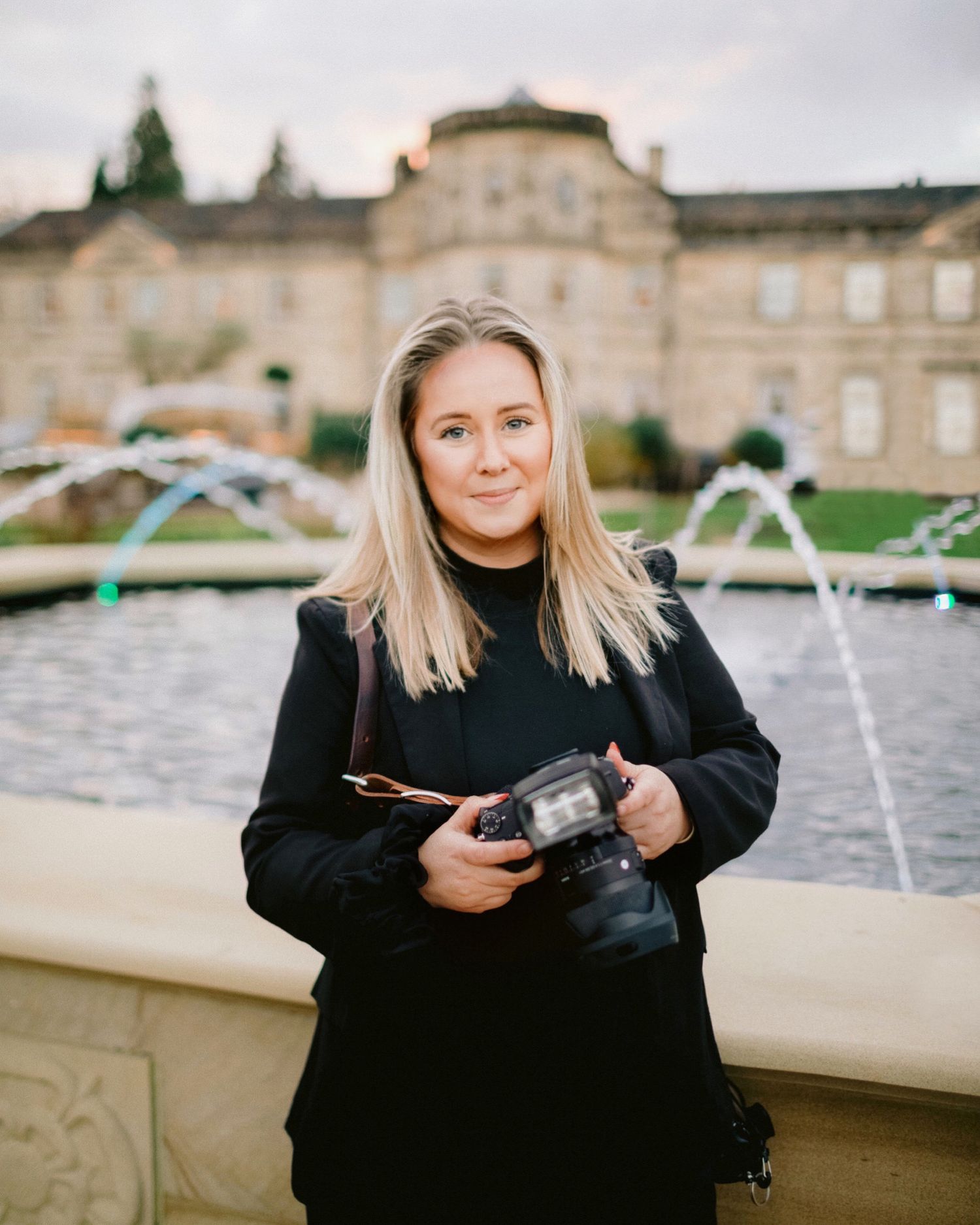 Female wedding photographer holding camera standing before a fountain at a stately venue.