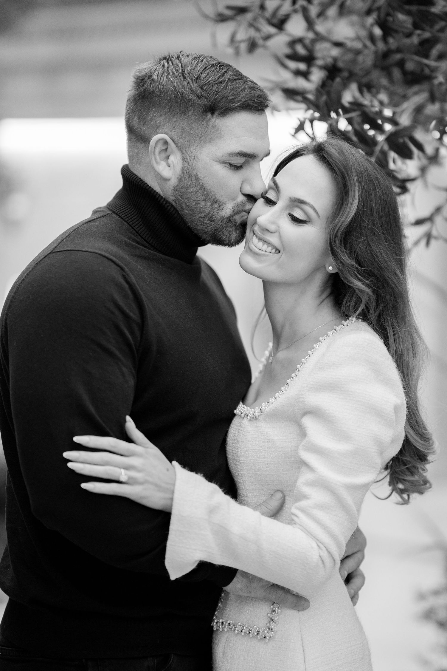 Black and white photo of couple sharing tender moment, man kissing smiling woman in white dress outdoors.
