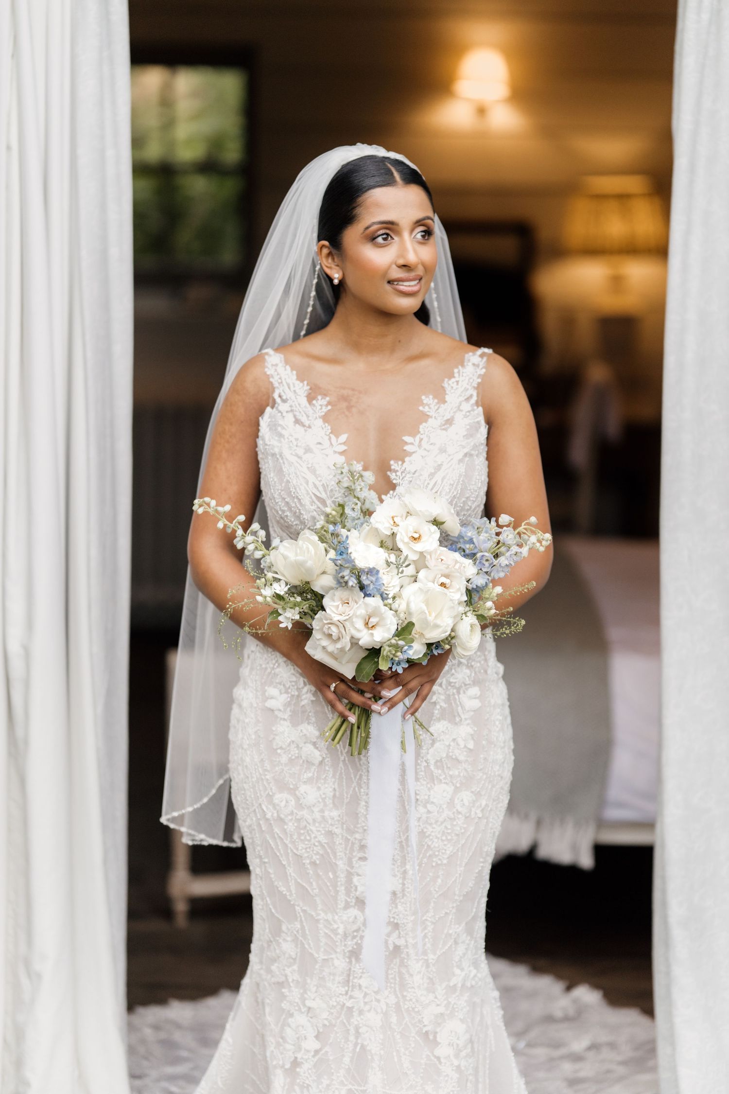 Bride in lace mermaid gown and veil holds white and blue bouquet, smiling elegantly in a softly lit doorway.