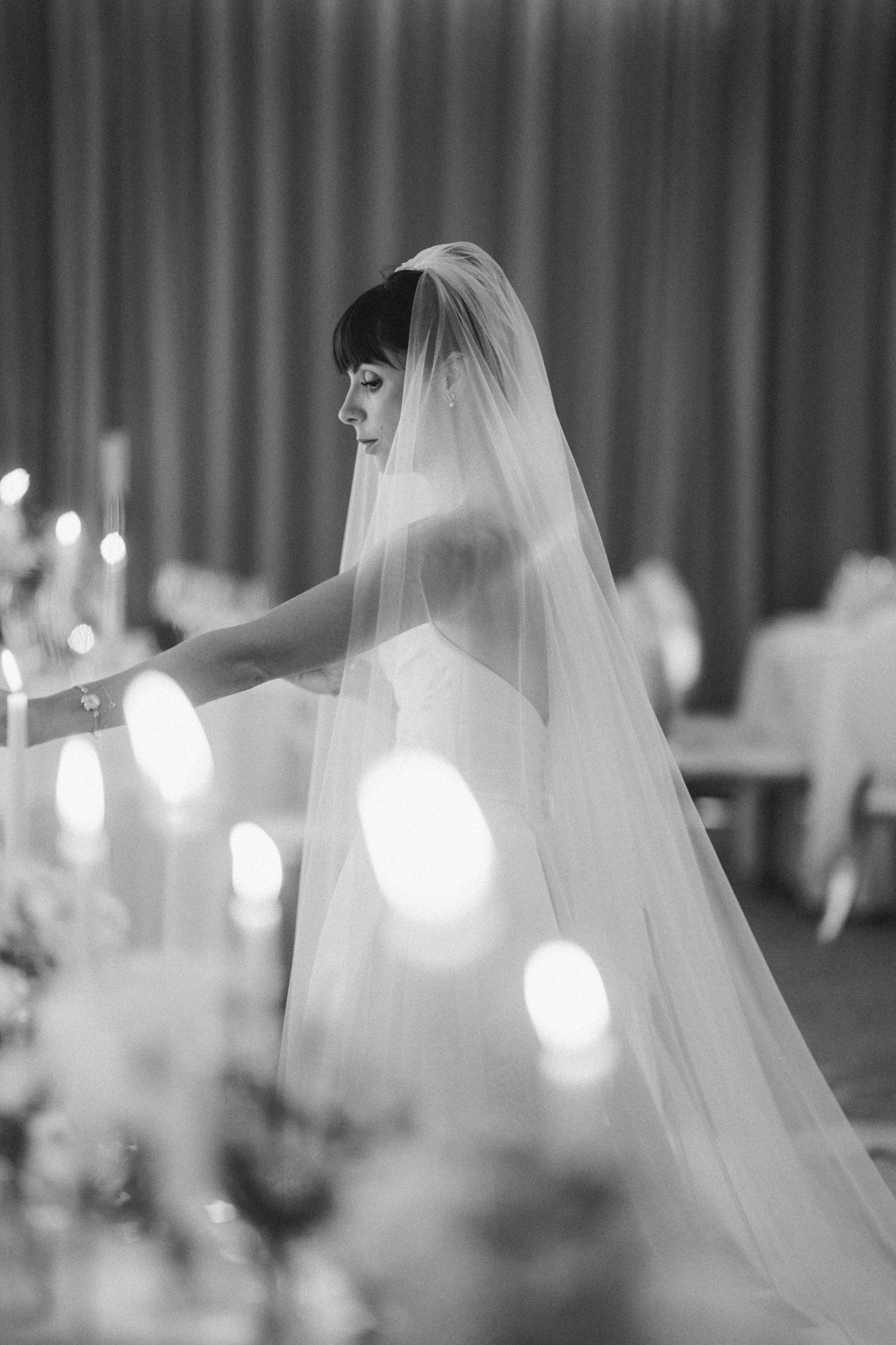 Black and white photo of a bride in a strapless gown and long veil standing among glowing candles.