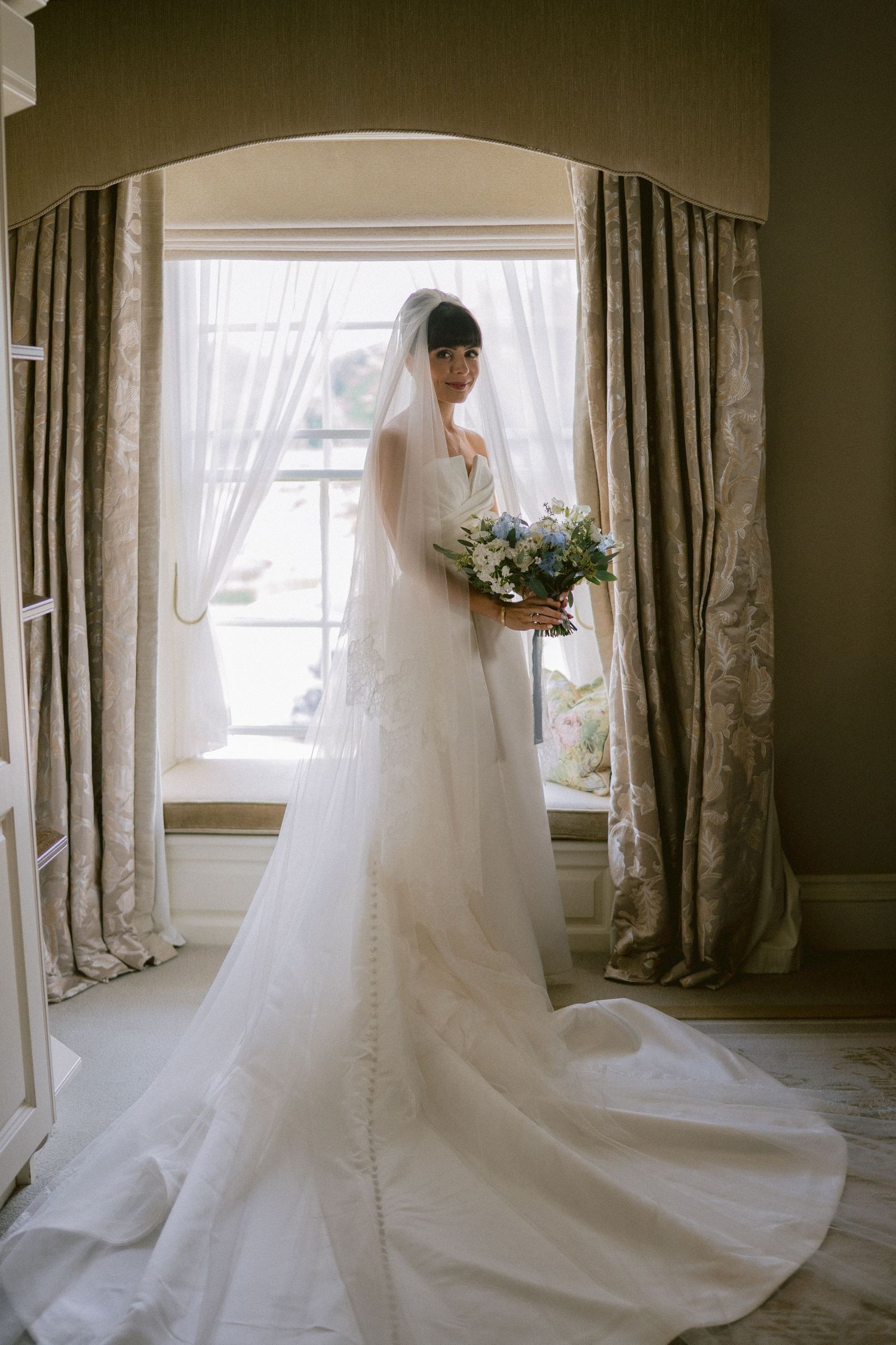 Bride in strapless lace wedding gown with cathedral veil holding blue and white bouquet by arched window.