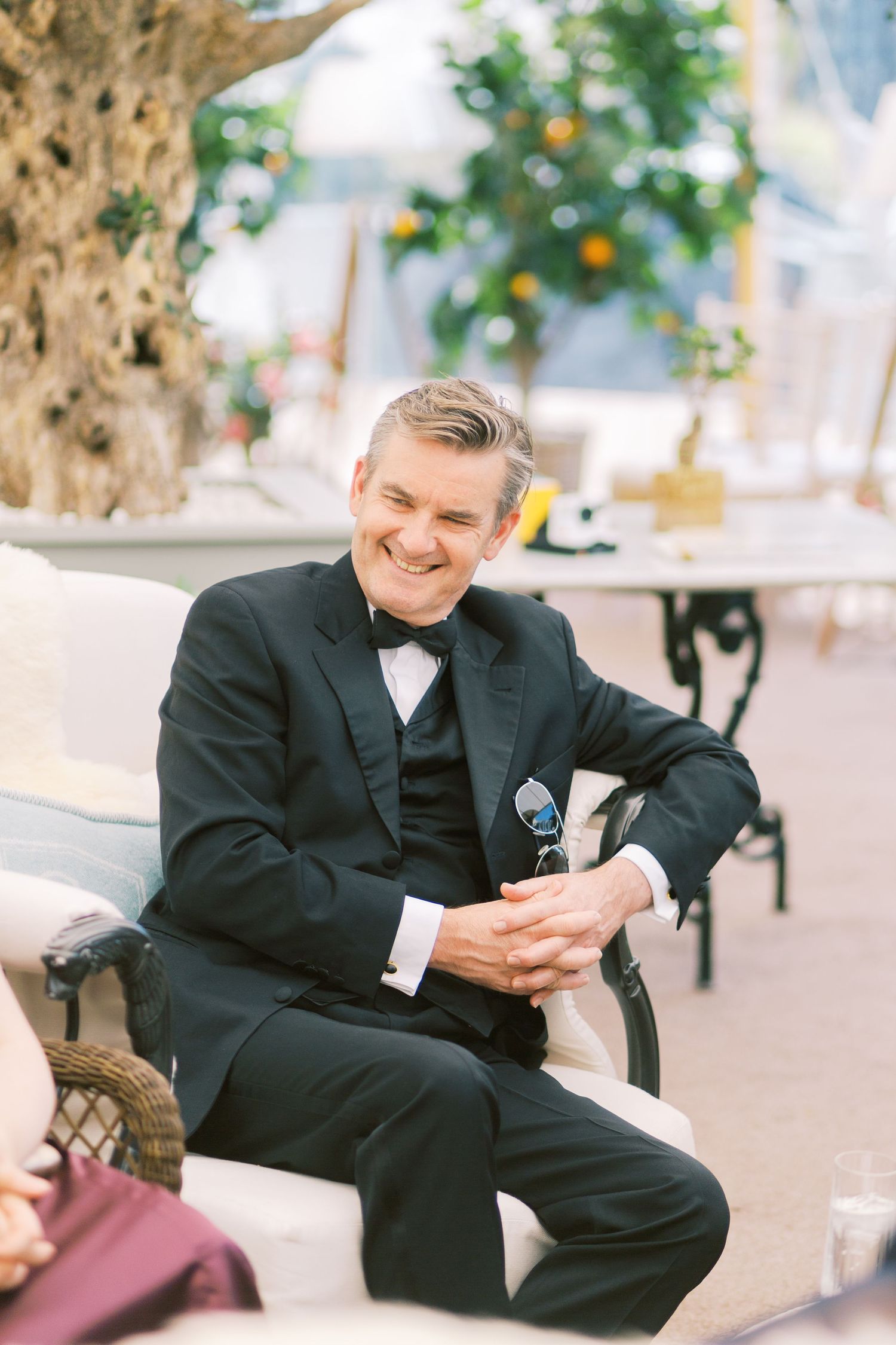 Smiling groom in black tuxedo and bow tie seated at outdoor wedding reception.