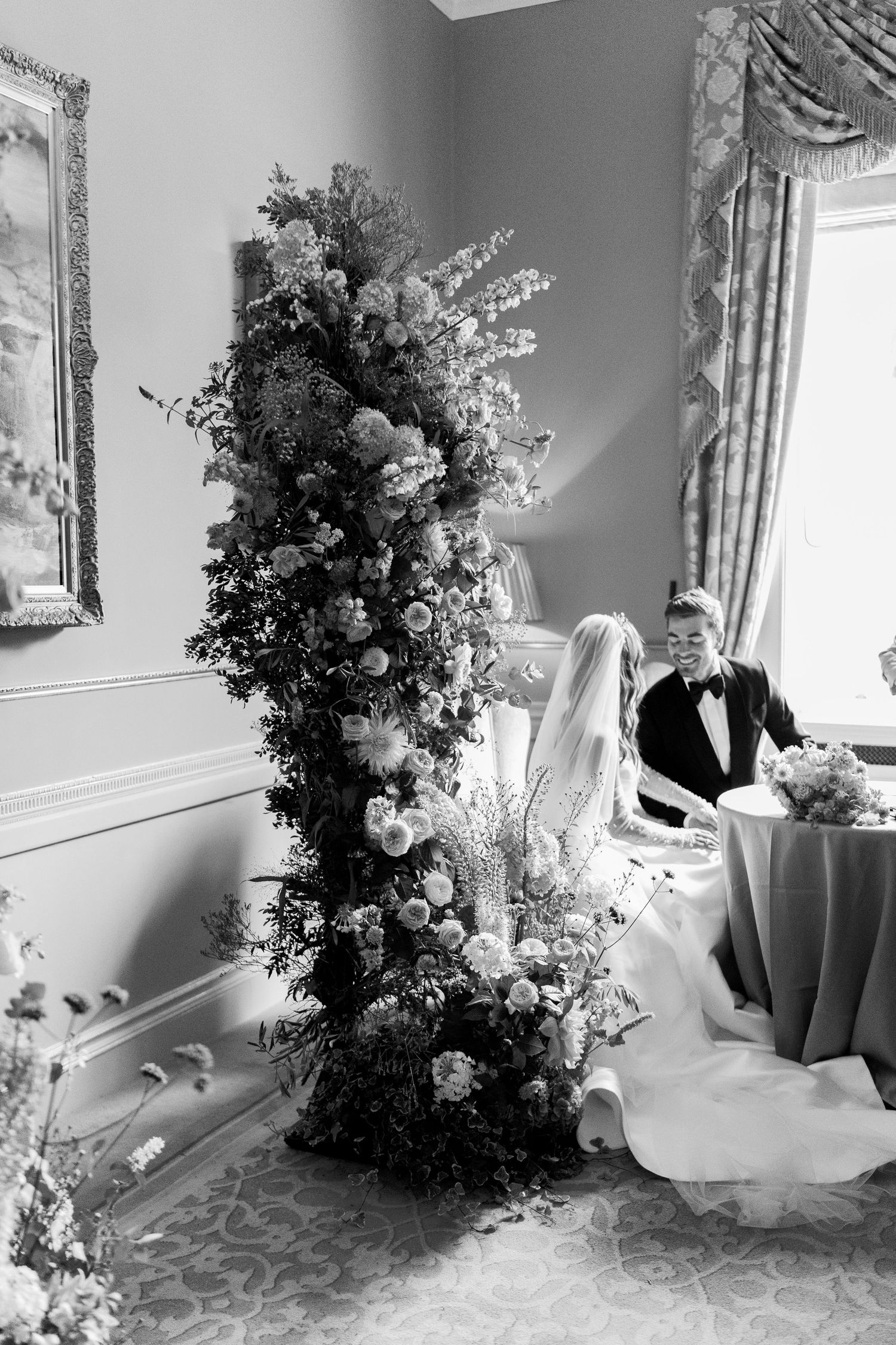 Bride and groom sit beside tall floral arrangement during intimate indoor wedding ceremony.