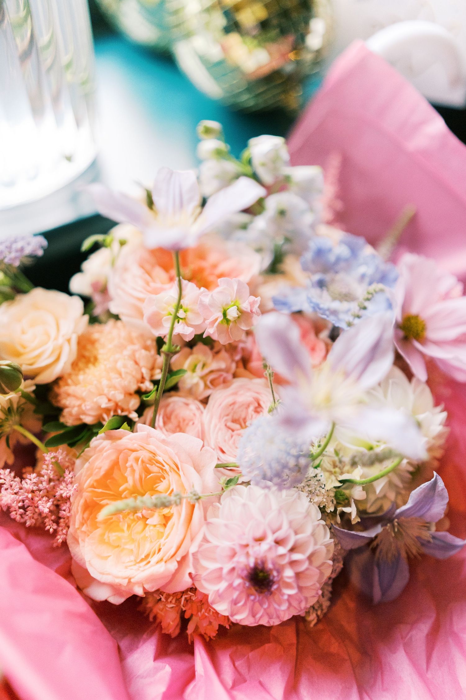 Romantic wedding bouquet featuring peach roses, pink dahlias, and blue wildflowers arranged on pink tissue paper.
