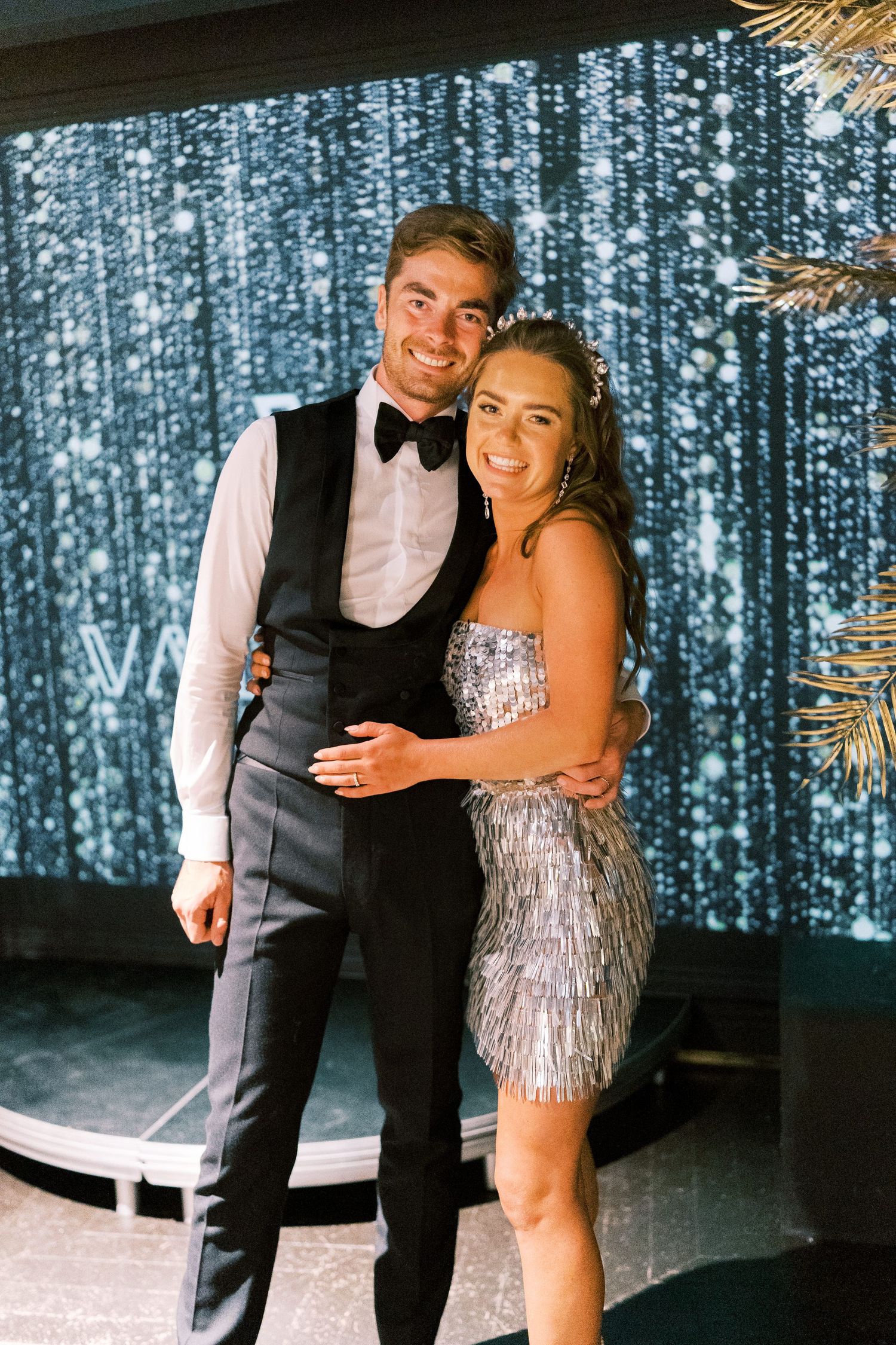 Young couple in formal attire posing together at a glamorous event, man in tuxedo vest and woman in silver sequin dress.