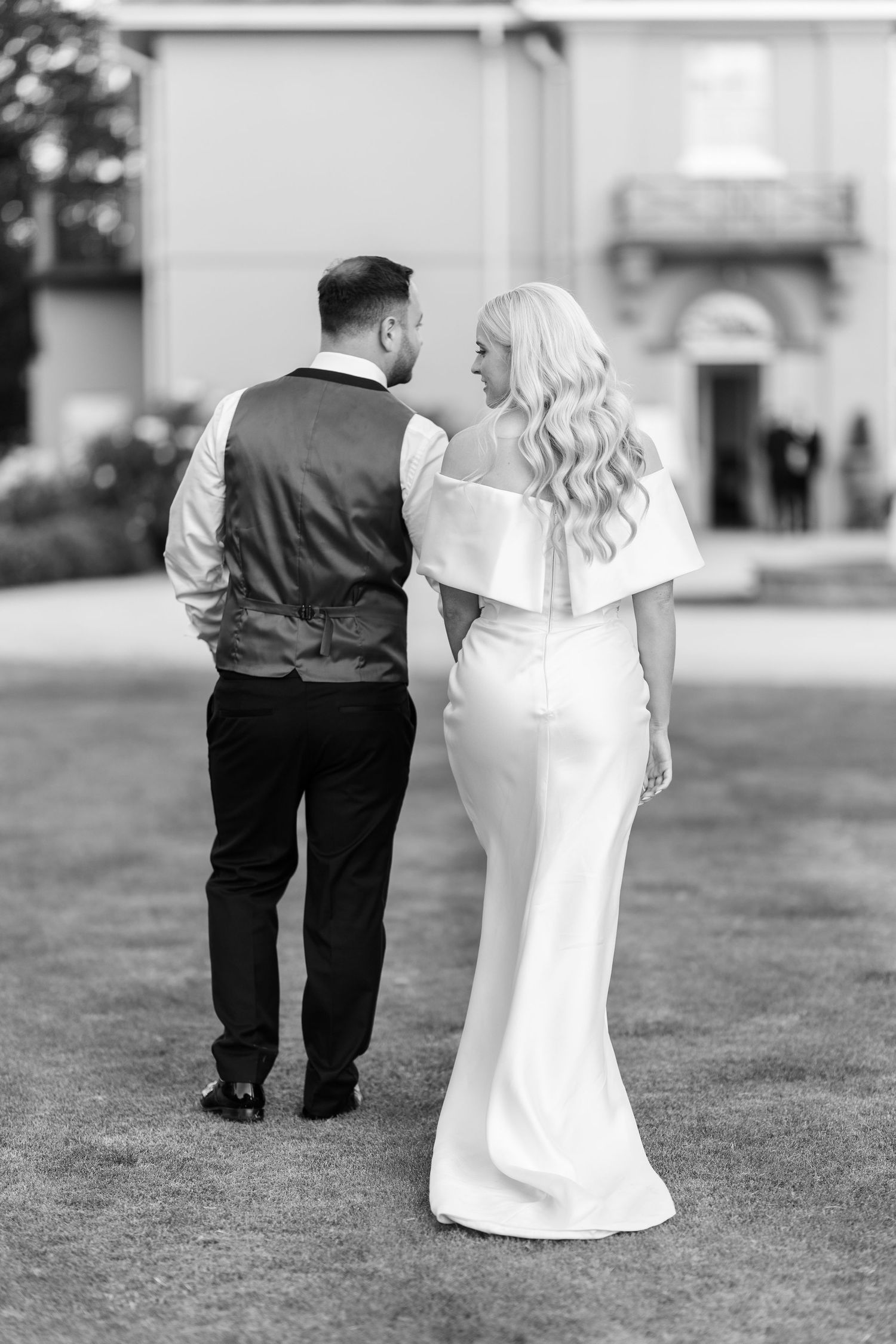Black and white rear view of bride and groom walking together toward an elegant venue.