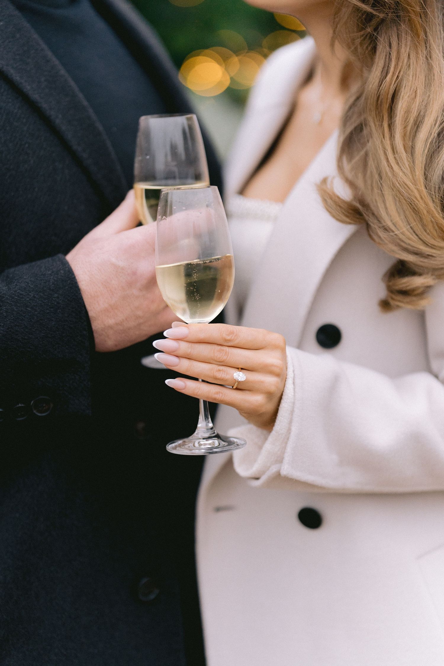 Close-up of engaged couple holding champagne glasses, showcasing diamond engagement ring on womans hand.