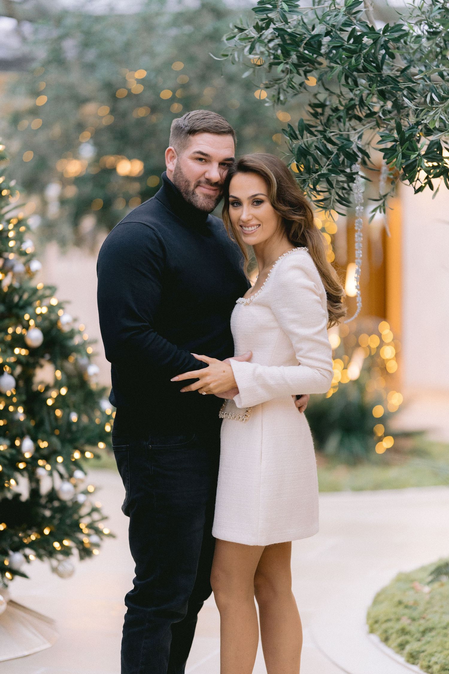 Happy couple embracing near decorated Christmas tree, man in black turtleneck and woman in white mini dress.